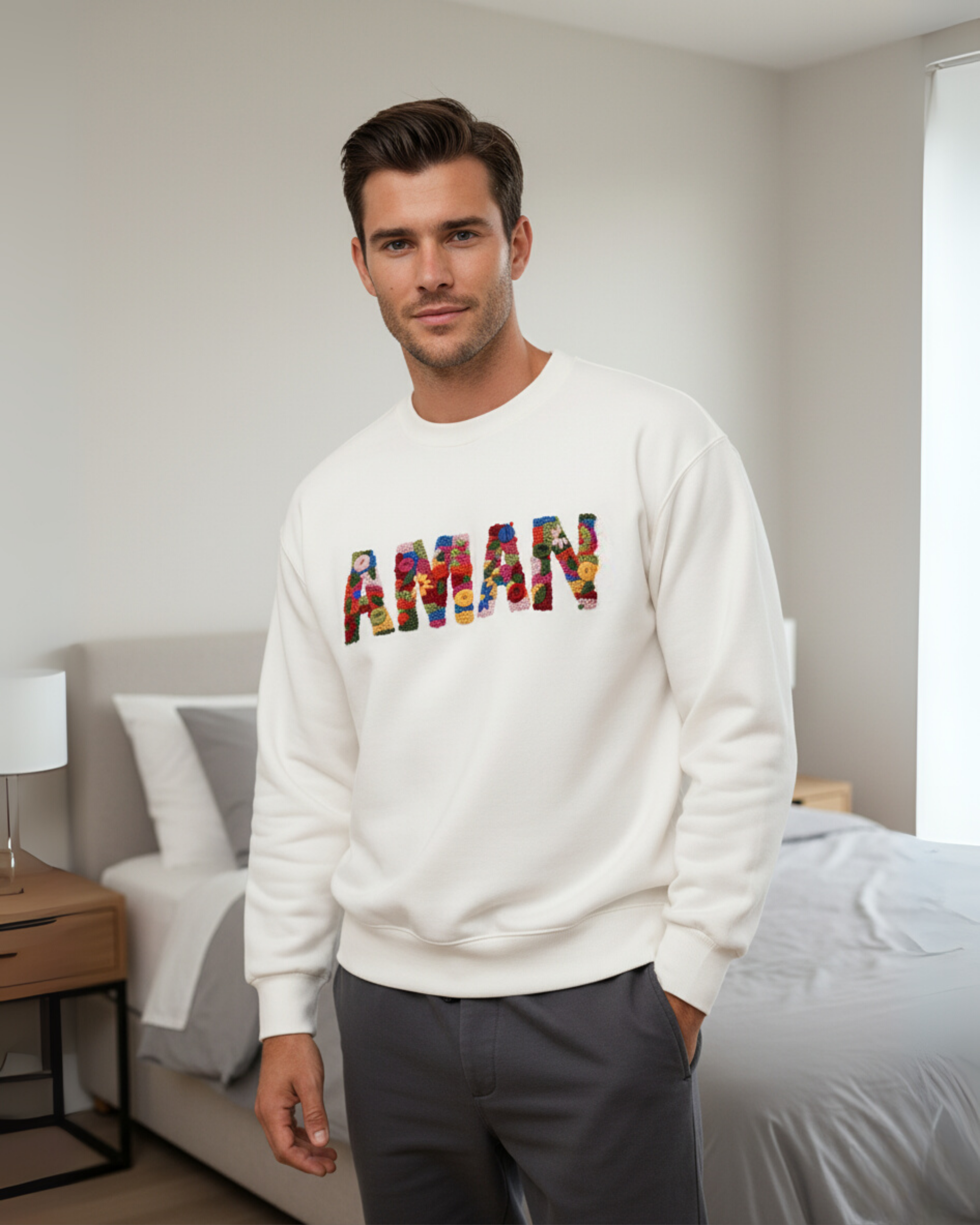 Man wearing a white sweatshirt with 'AMAN' embroidered in colorful letters in a bedroom setting.