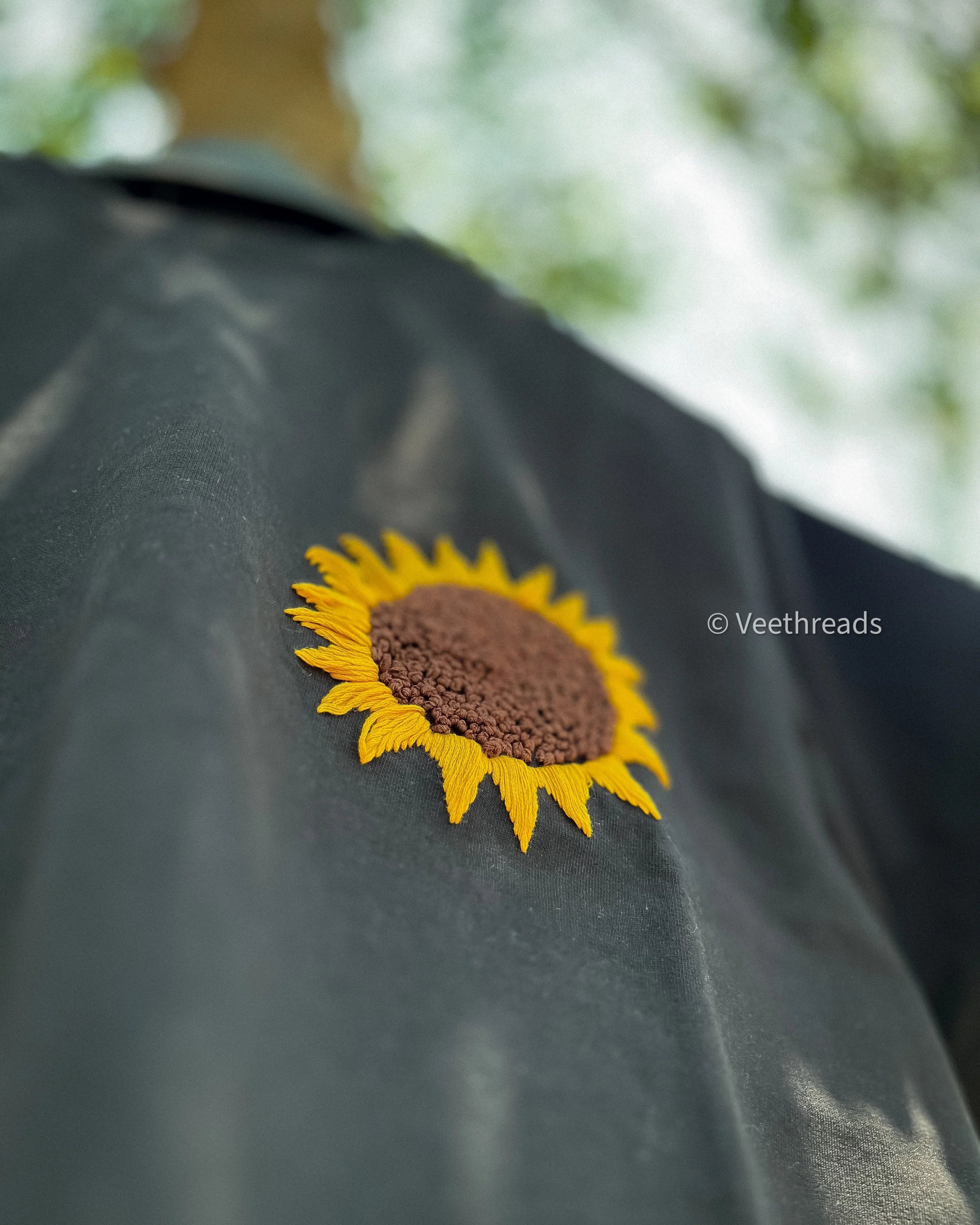 Close-up of a hand-embroidered yellow sunflower on a black cotton tee showing raised real thread