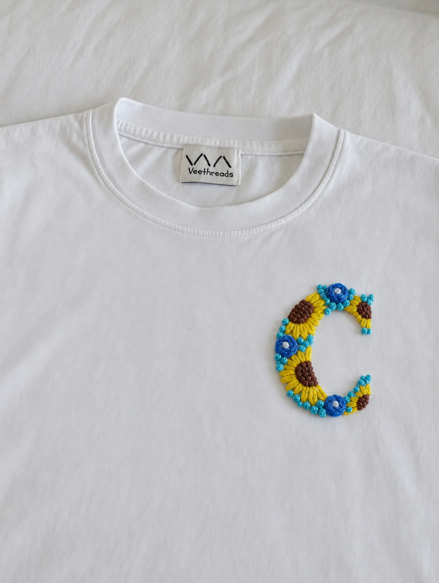 Sunflower and blue-flower hand-embroidered letter on a white cotton tee, by Vee Threads