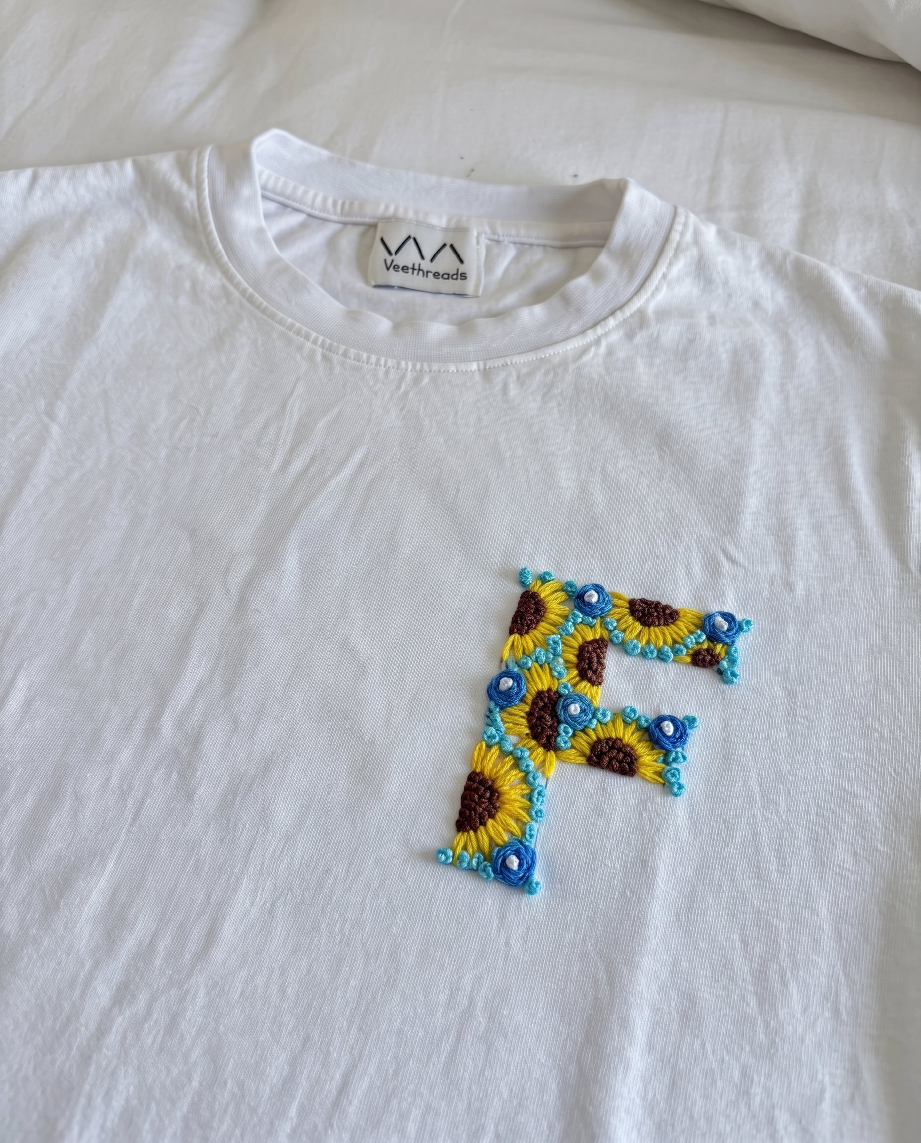 White cotton tee with an initial hand-embroidered as sunflowers and small blue flowers, by Vee Threads