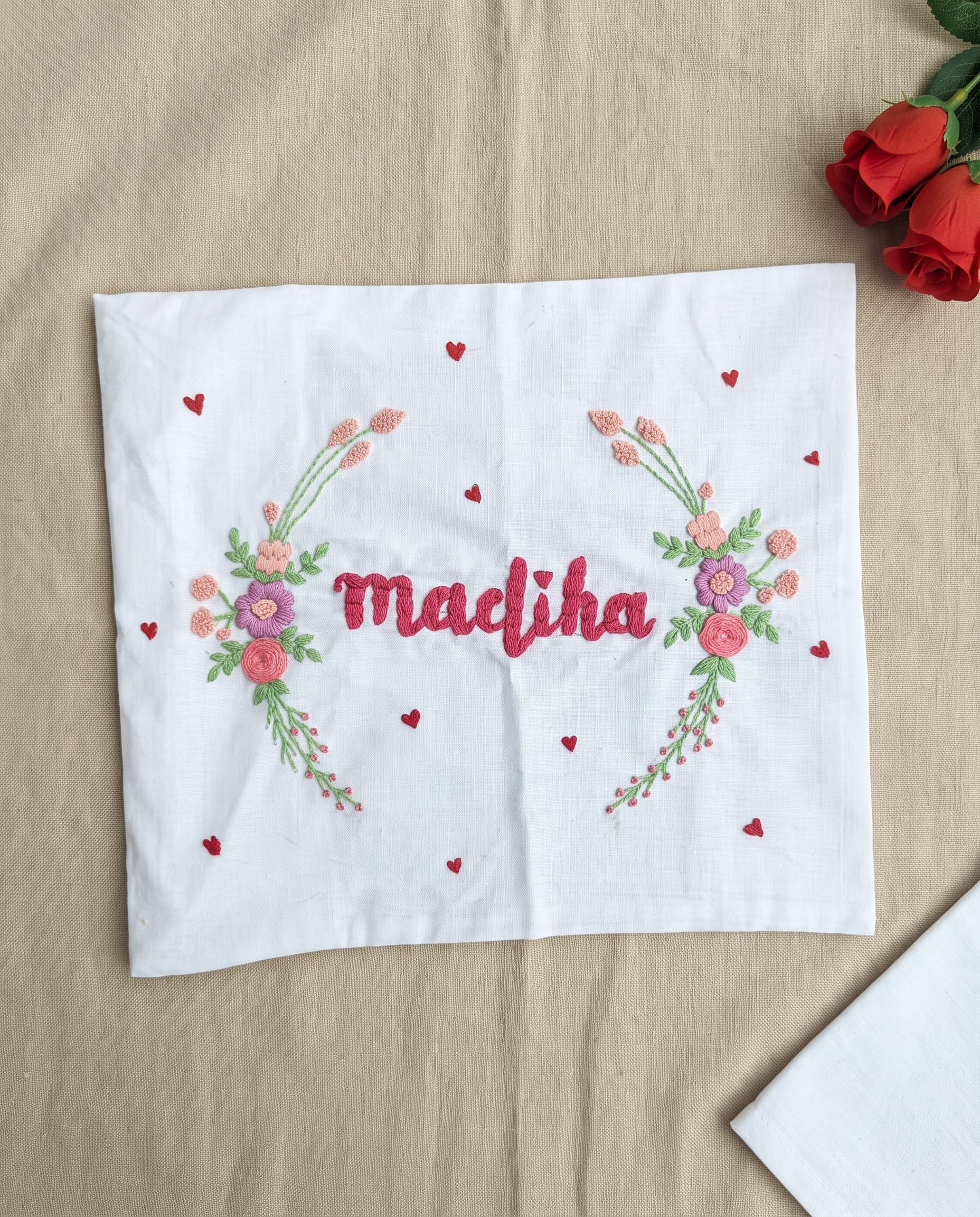 A white embroidered cloth napkin or handkerchief laid flat on a beige fabric surface, featuring the name 'Madhika' in red script between two floral garlands with pink, red, green, and purple flowers and small scattered red hearts. Red roses appear in the top-right corner, and part of another white cloth is visible in the bottom-right.