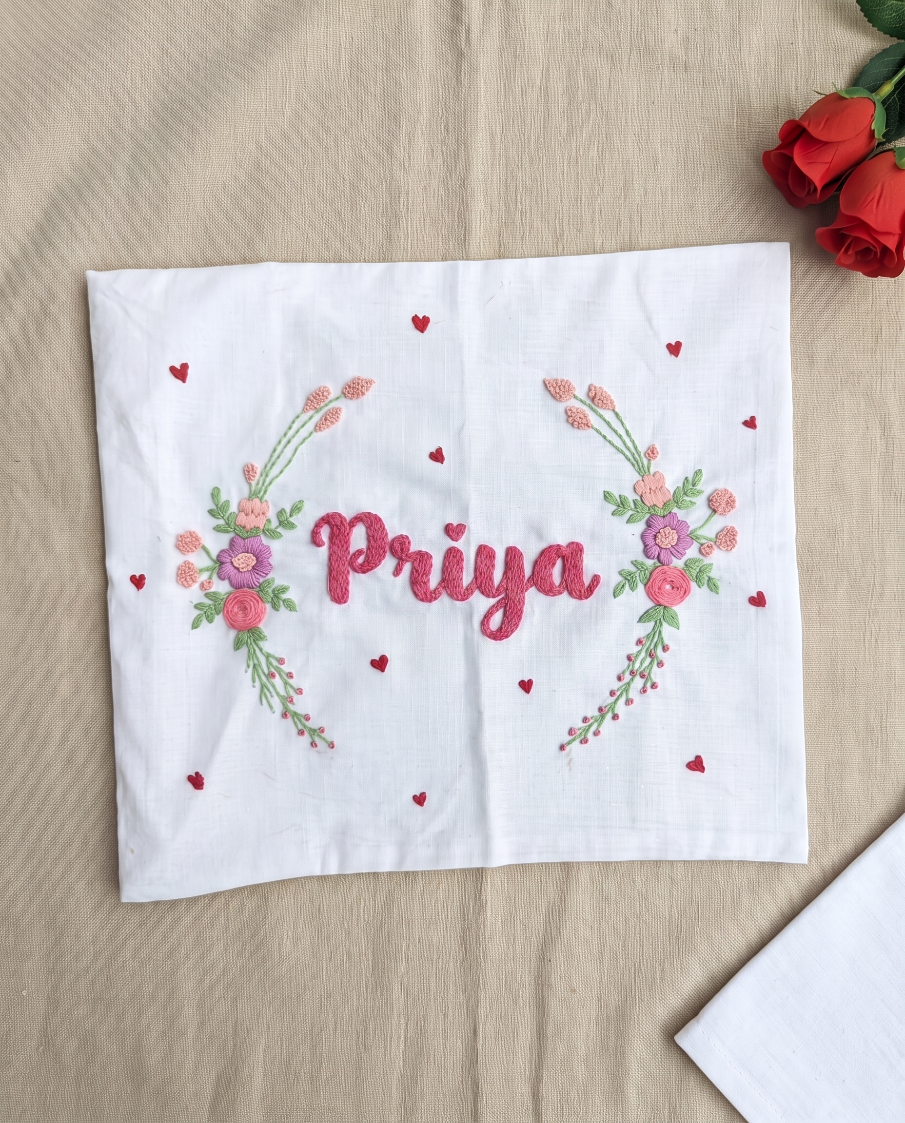 A white embroidered handkerchief custom the name 'Priya' in pink script, flanked by pink and purple floral garlands with small red hearts, laid flat on a beige fabric surface with red roses in the corner.