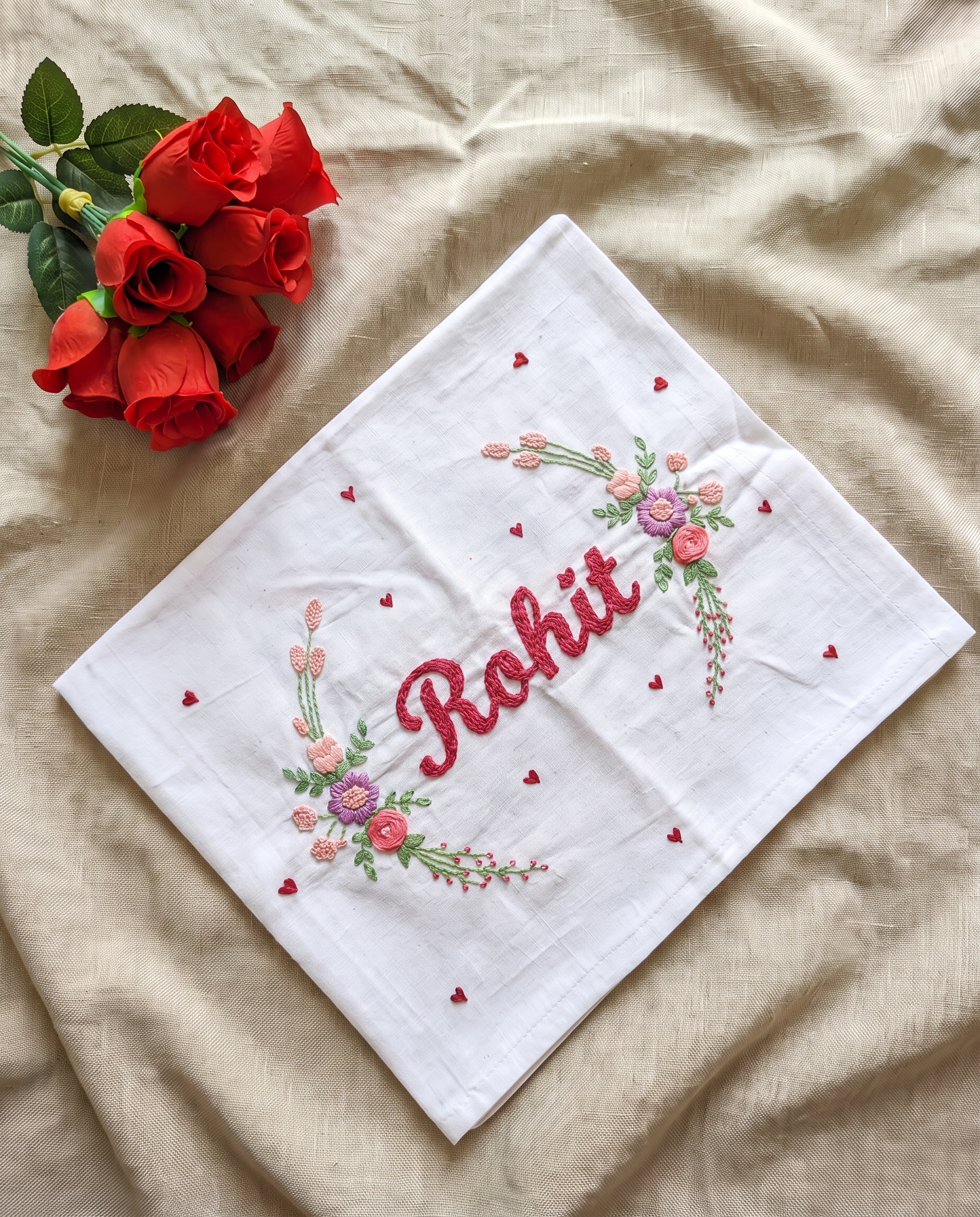 A white embroidered hanky  with the name 'Rohit' stitched in red script, surrounded by pink and green floral motifs and small red hearts, laid flat on beige fabric next to red roses.