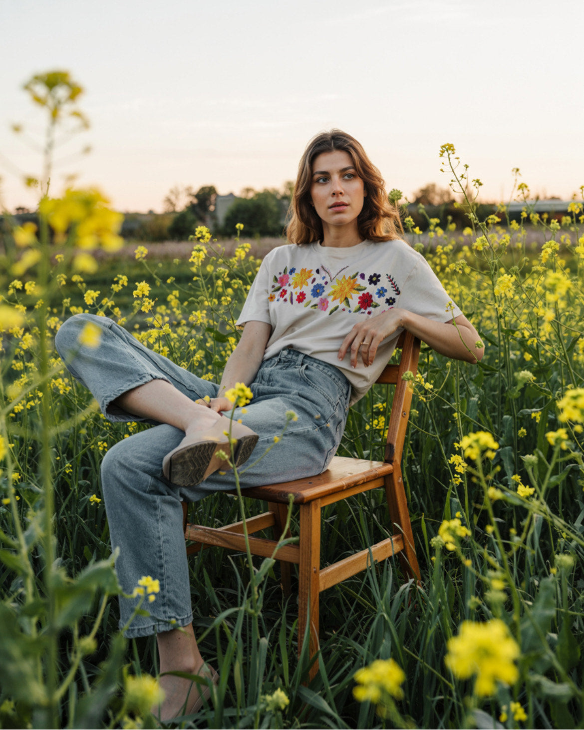 A person sitting on a chair in a field of yellow flowers, wearing a white oversized t-shirt with vibrant floral embroidery and blue denim jeans.