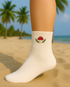White sock with a mushroom embroidery design on a sandy beach with palm trees and ocean in the background