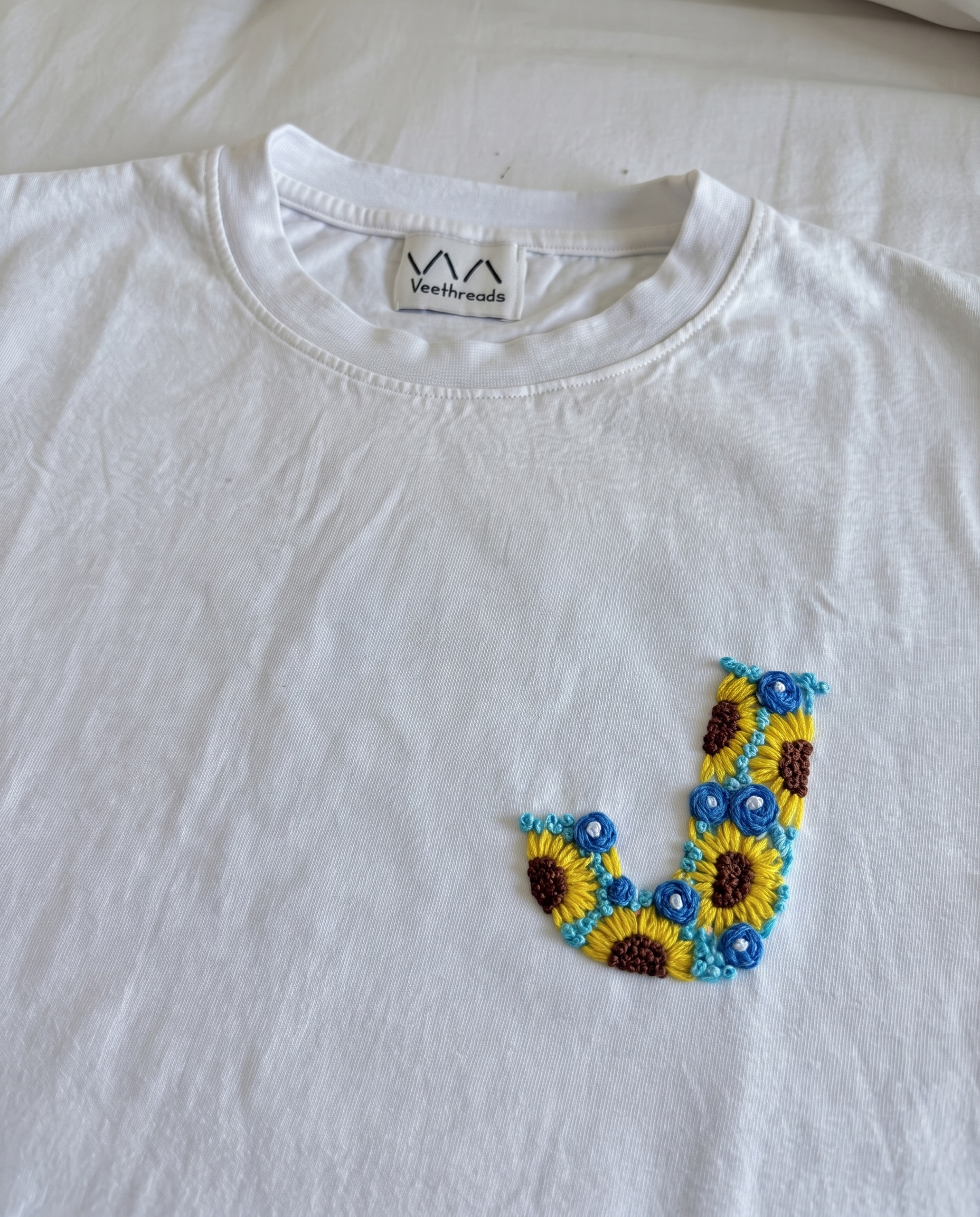 White cotton tee with an initial hand-embroidered as sunflowers and small blue flowers, by Vee Threads