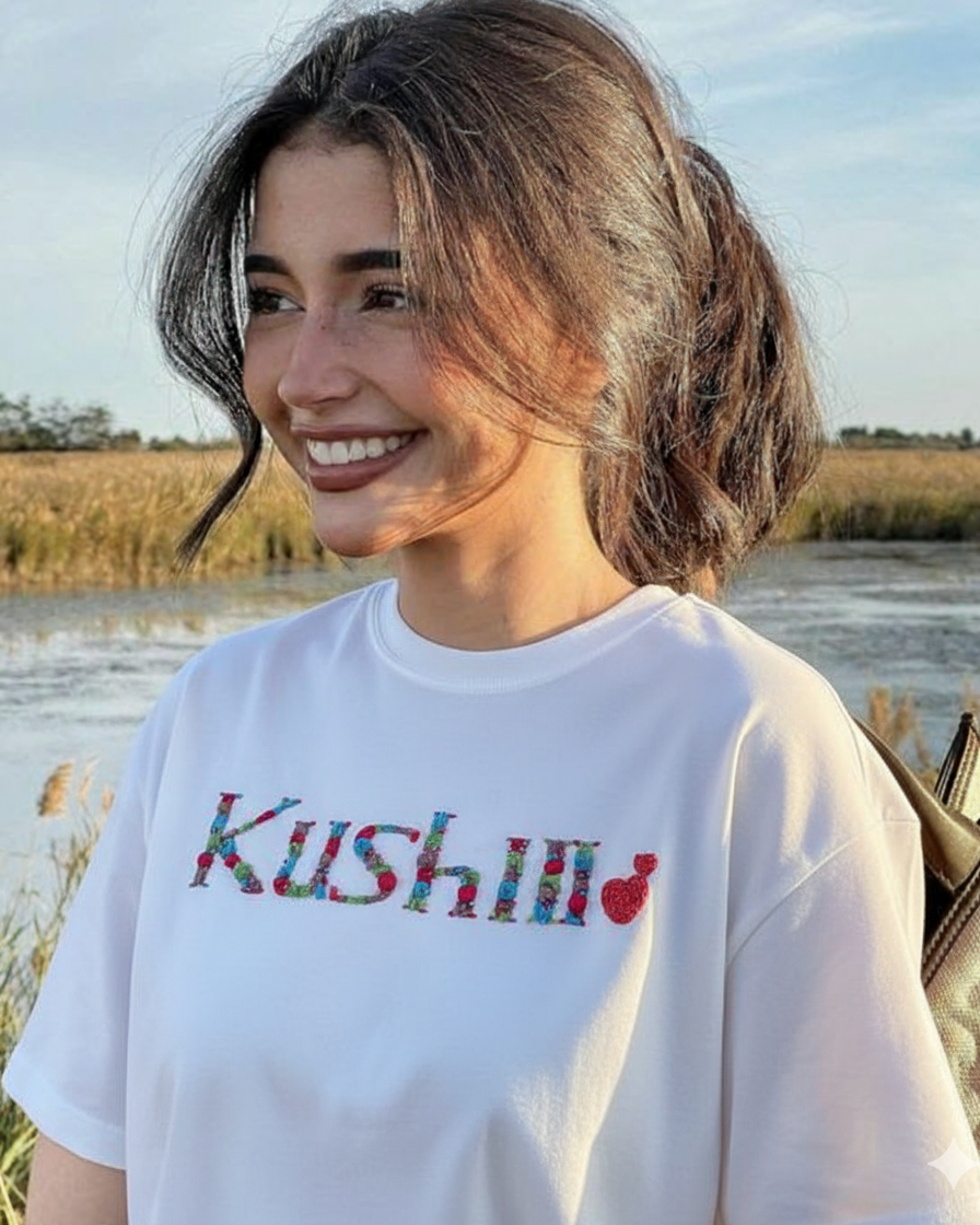 Woman wearing a white t-shirt with 'Kushii' name embroidered  on it, standing outdoors by a body of water.