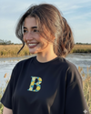 Woman wearing a black t-shirt with a colorful 'B' letter name custom embroidered , standing outdoors by a body of water.