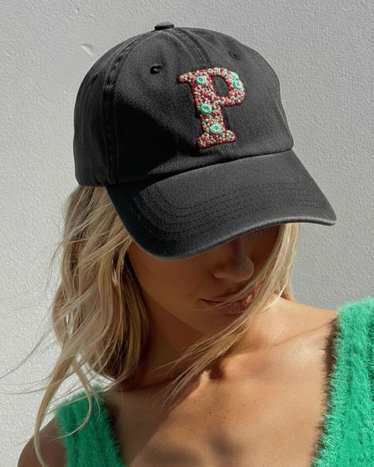 Black cap with a decorative 'P' initial embroidered worn by a person in a green top against a gray background