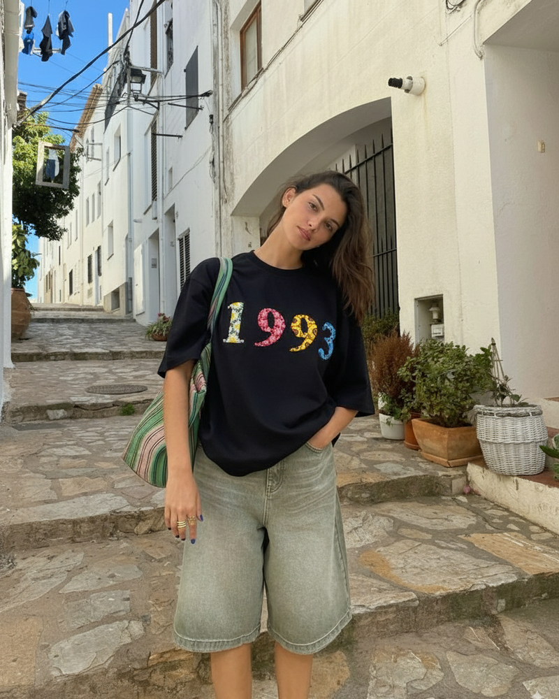 Woman wearing a black t-shirt with '1993' custom year embroidered  on it, standing on a stone street.
