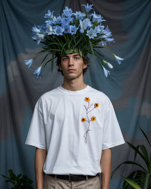 A young man with light skin and dark hair wears a white t-shirt adorned with embroidered blue flowers and green leaves. He also wears an elaborate floral headpiece made of real blue flowers extending outward like a crown. His expression is calm and serious, with a silver chain necklace adding a subtle touch. The background features lush blue flowers and green foliage, enhancing the surreal and artistic atmosphere of the scene. The overall aesthetic is ethereal and fashion-forward, blending nature with conte