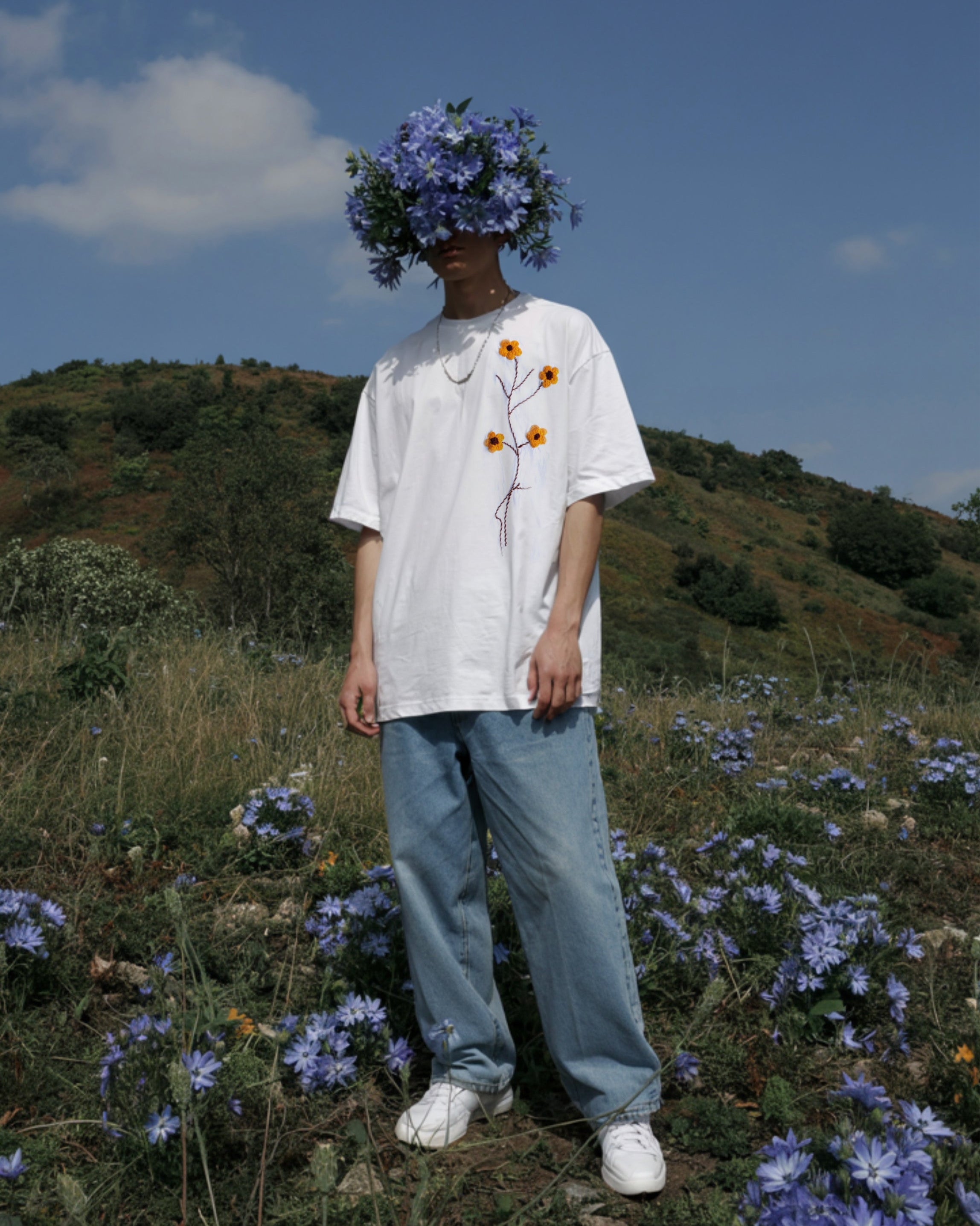 A young man with a serene expression stands in an artistic setting, wearing a white t-shirt embroidered with delicate blue flowers and green leaves. His striking floral headpiece, composed of real blue flowers, extends outward like a blooming halo. The backdrop is filled with lush blue blossoms and greenery, creating a dreamy, surreal atmosphere that merges fashion with nature.