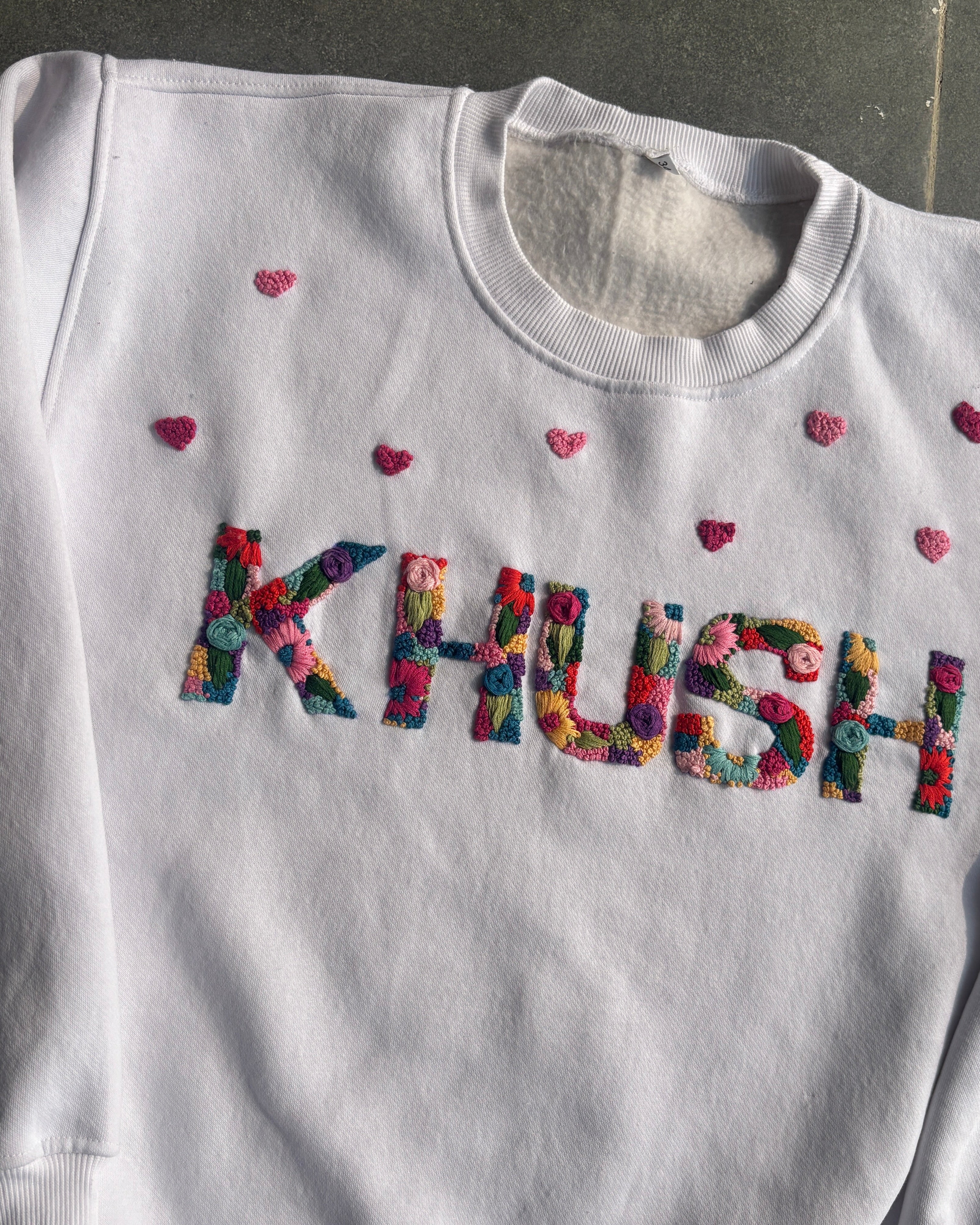 White sweatshirt with colorful 'KHUSH' customised  name hand embroidered  and small red hearts on a dark background