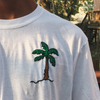White t-shirt with a palm tree design on the sleeve, outdoors.