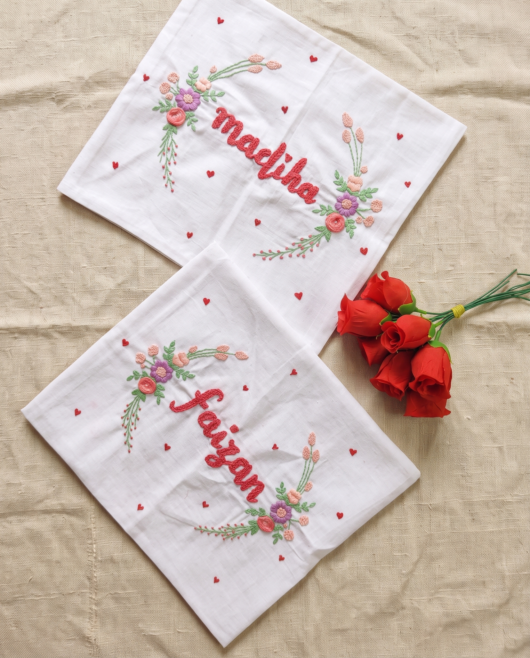 Two white embroidered cotton handkerchief with floral designs and personalized names 'madiba' and 'faizan', arranged on a beige fabric background next to red roses.