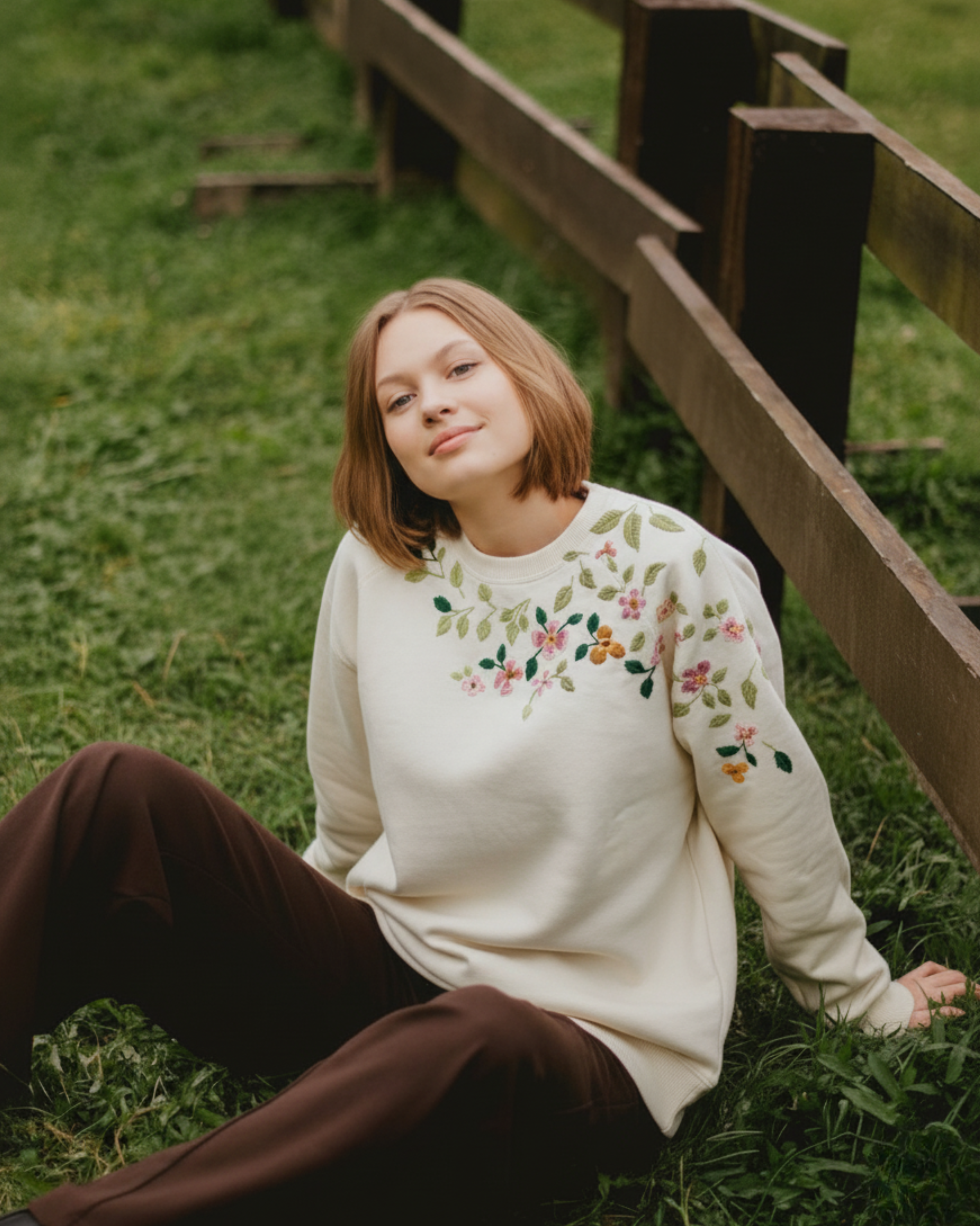 Woman wearing a cream sweater with floral embroidery sitting on grass next to a wooden fence.