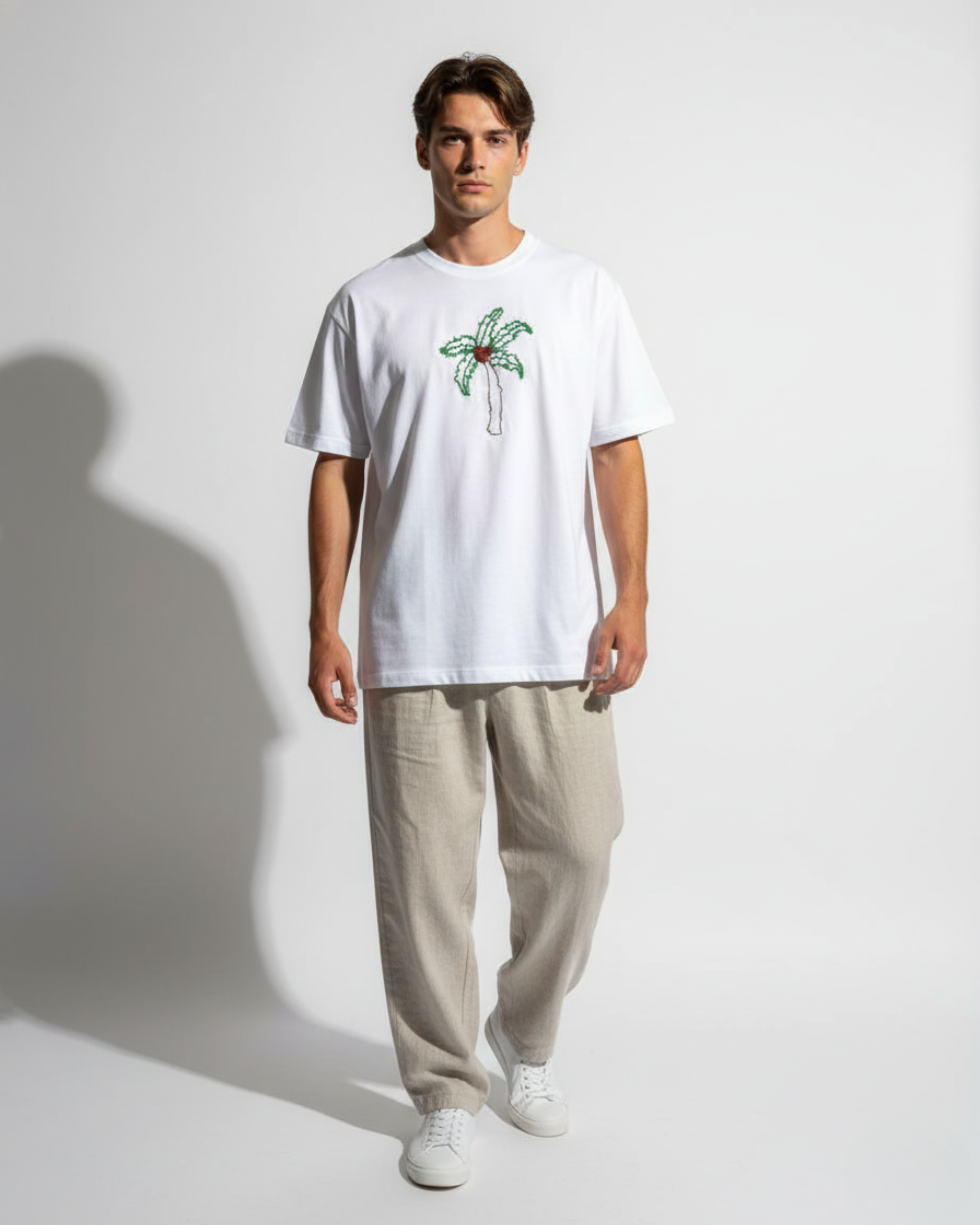 Man wearing a white t-shirt with a palm tree design bead embroidered and beige pants on a plain background