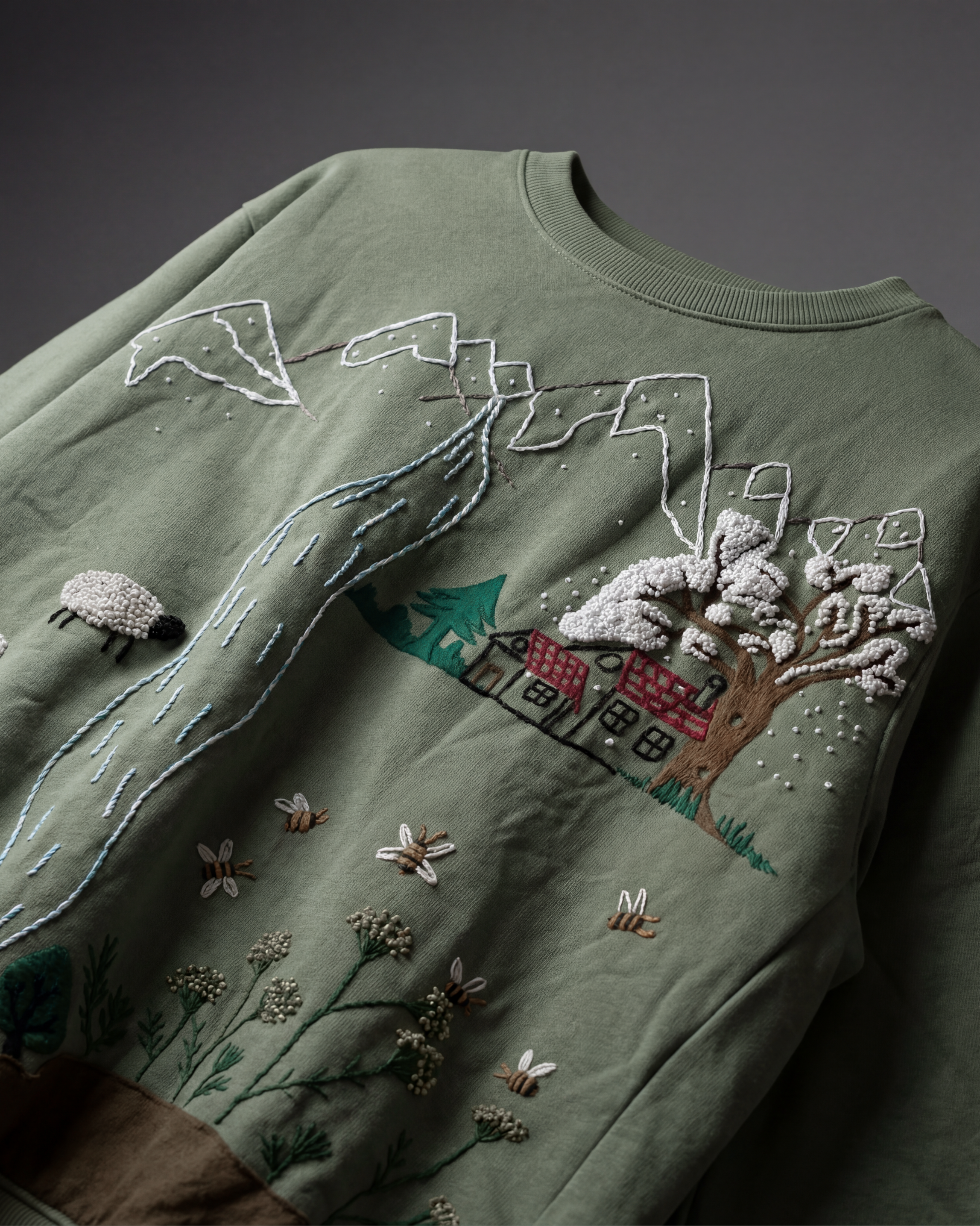 Green sweater with embroidered and hand painted landscape design featuring trees, a house, and animals on a dark background.