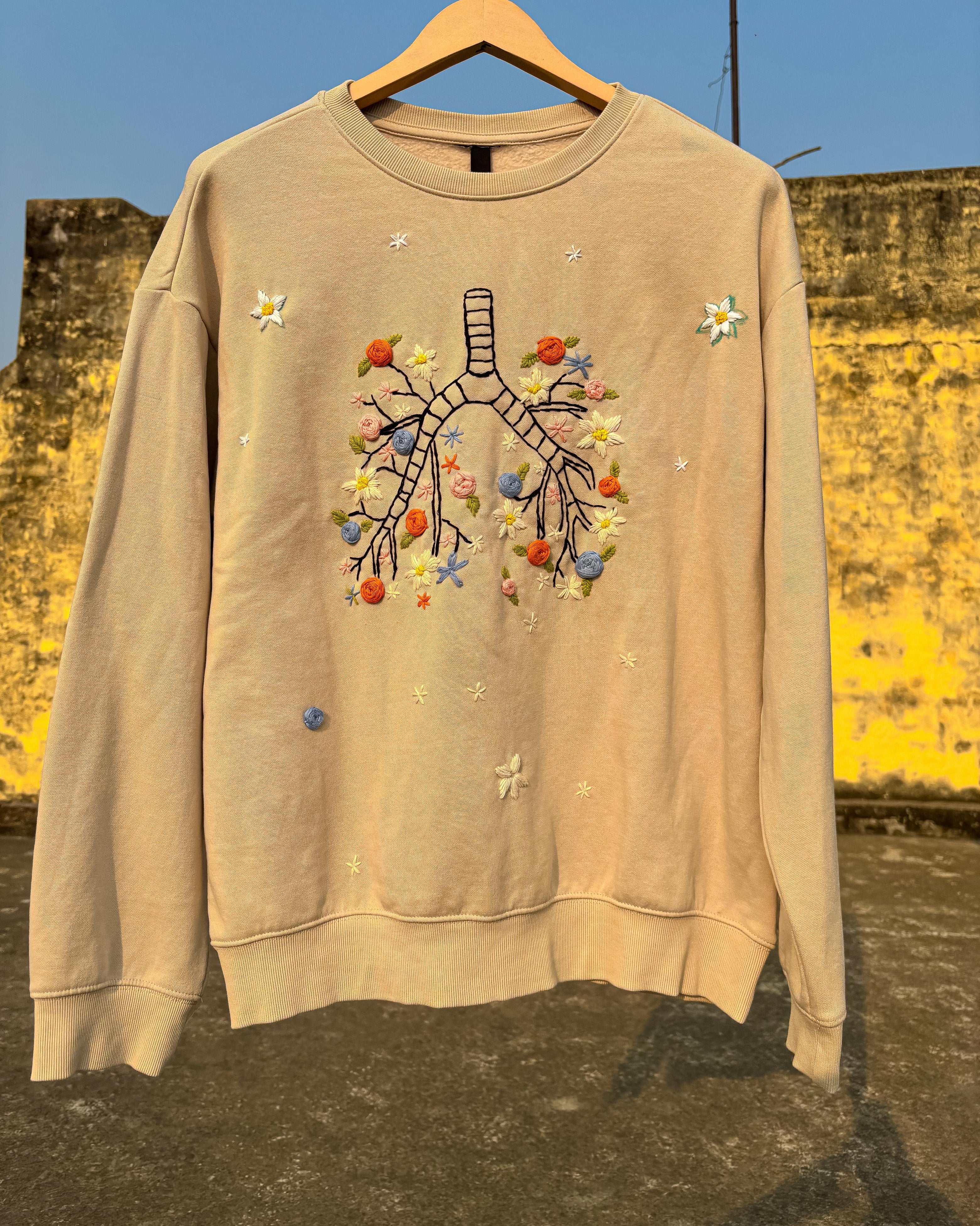 Beige sweatshirt with floral and abstract design on a wooden hanger against a yellow wall.