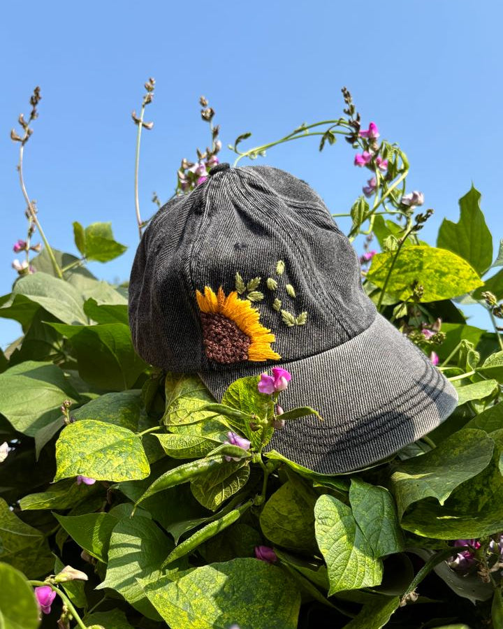 Gray cap with embroidered sunflower design on a plant with green leaves and small flowers.