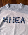 White sweatshirt with 'RHEA' in floral embroidered letters on a wooden surface