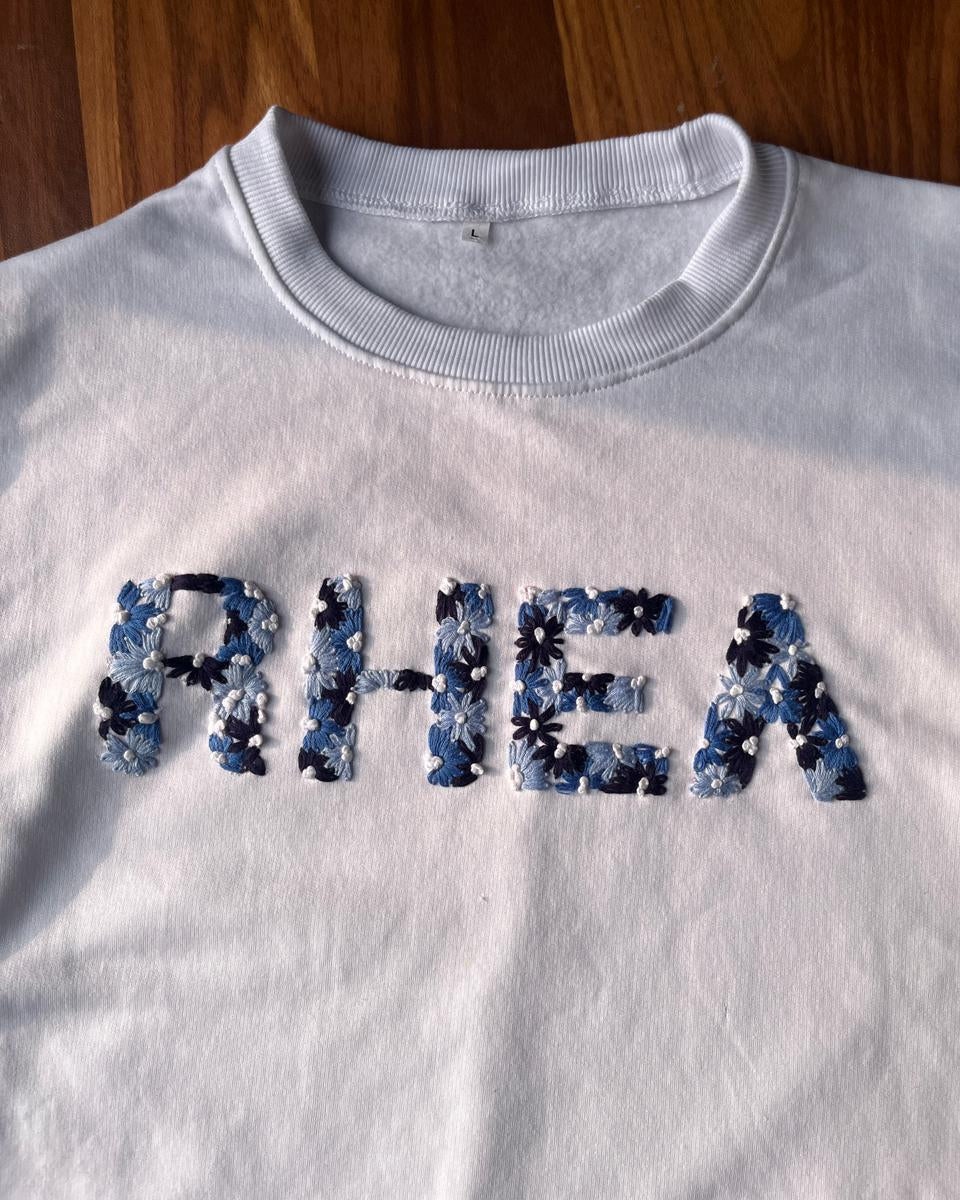 White sweatshirt with 'RHEA' in floral embroidered letters on a wooden surface