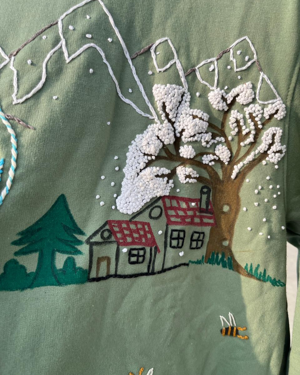 Embroidered design of a snowy landscape with trees and houses on a green fabric.