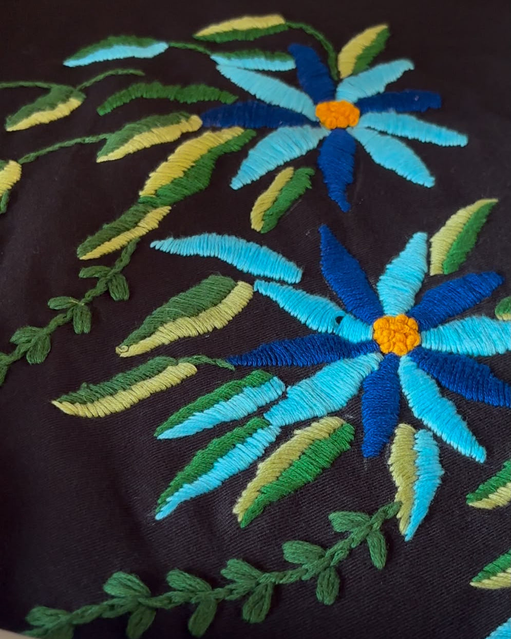 Close-up of embroidered floral pattern on a black tshirt, featuring blue and light blue flowers with yellow centers and green and yellow leaves.