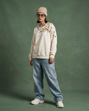 Person wearing a cream sweater with floral embroidery, light blue jeans, and white sneakers on a green background