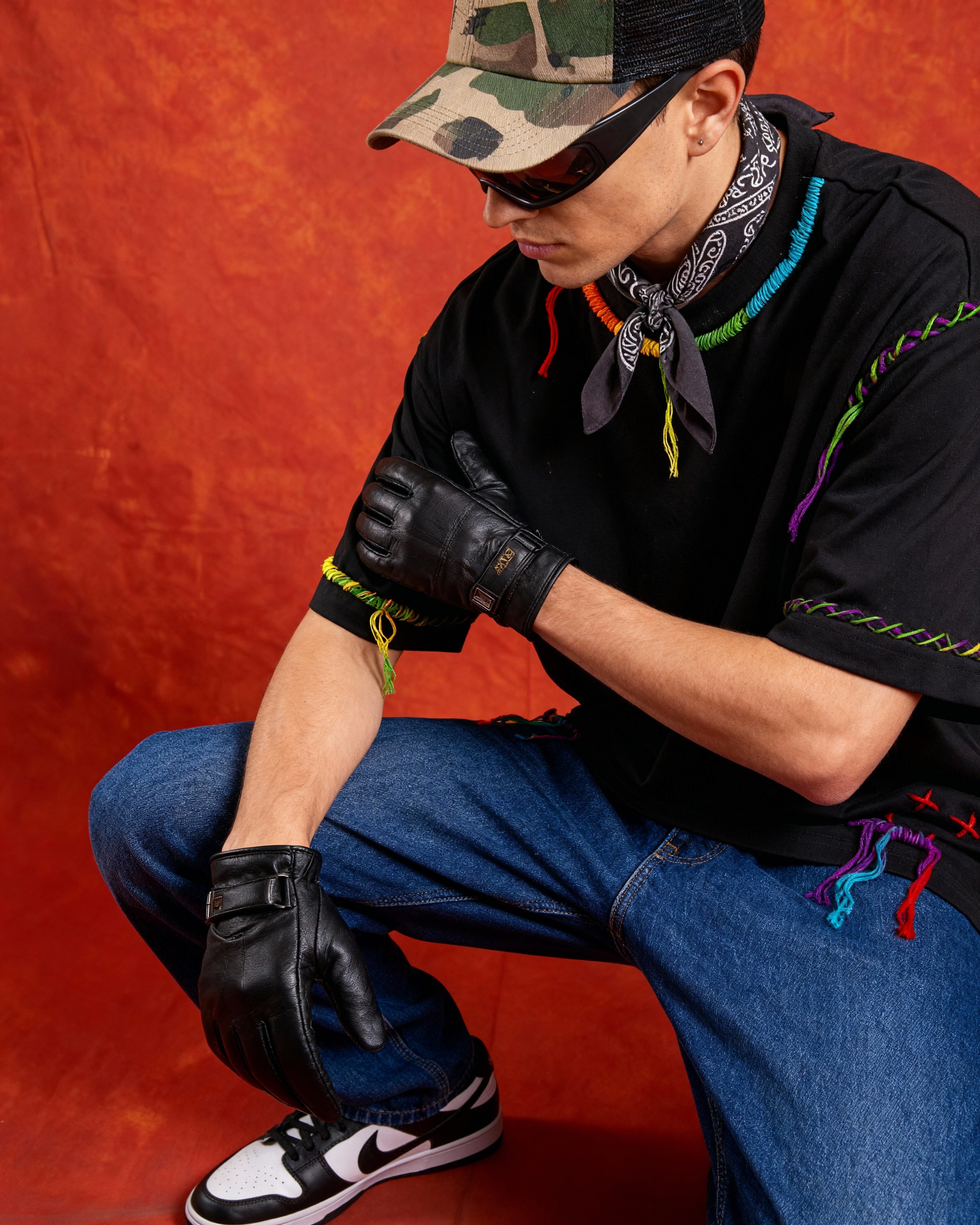 Person wearing a black shirt with colorful embroidered accents, blue jeans, black gloves, and a camouflage cap on a red background.