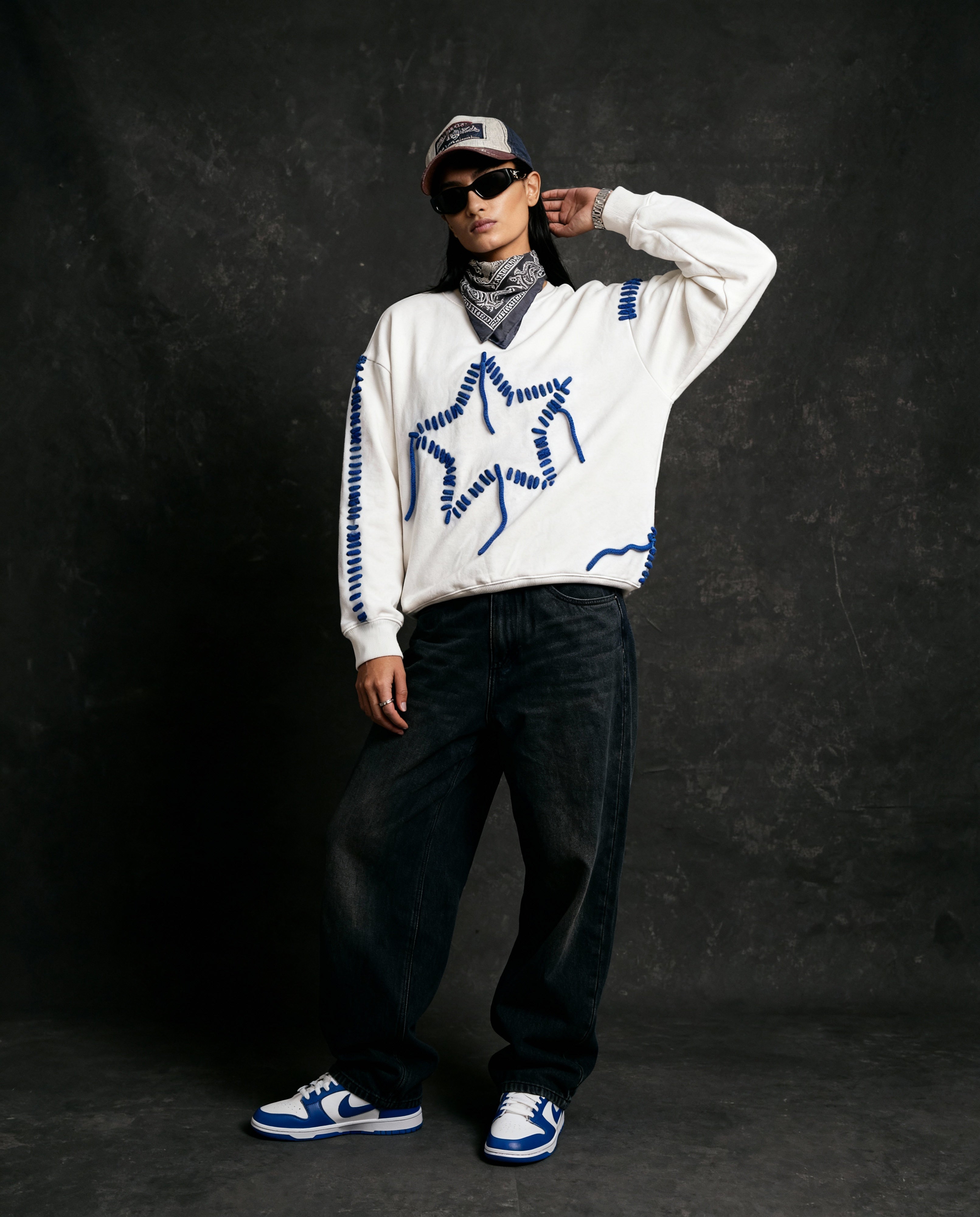 Person wearing a white sweater with blue patterns, dark pants, and sneakers on a black background