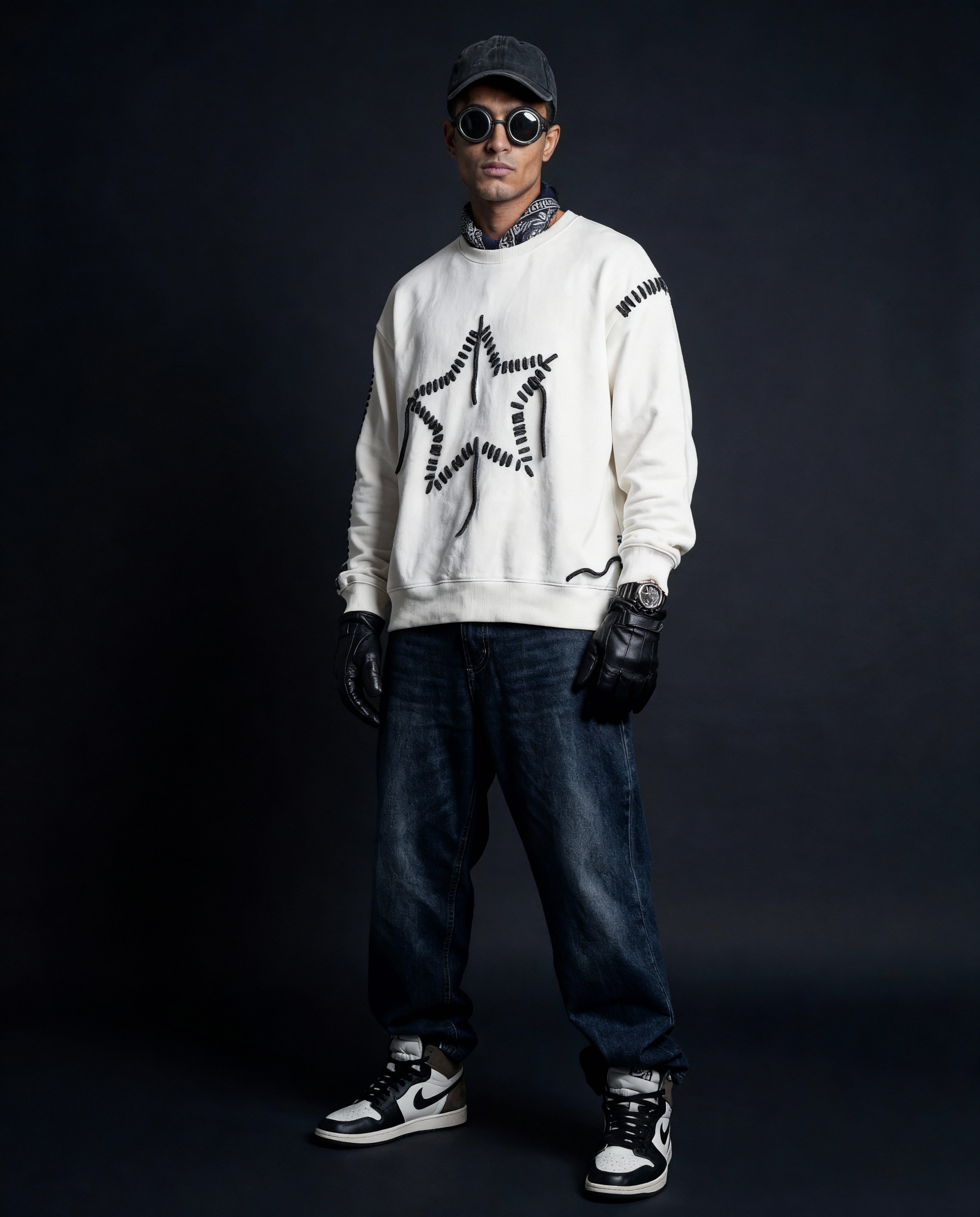 Person wearing a white sweatshirt with black designs, dark jeans, black gloves, and a cap on a dark background