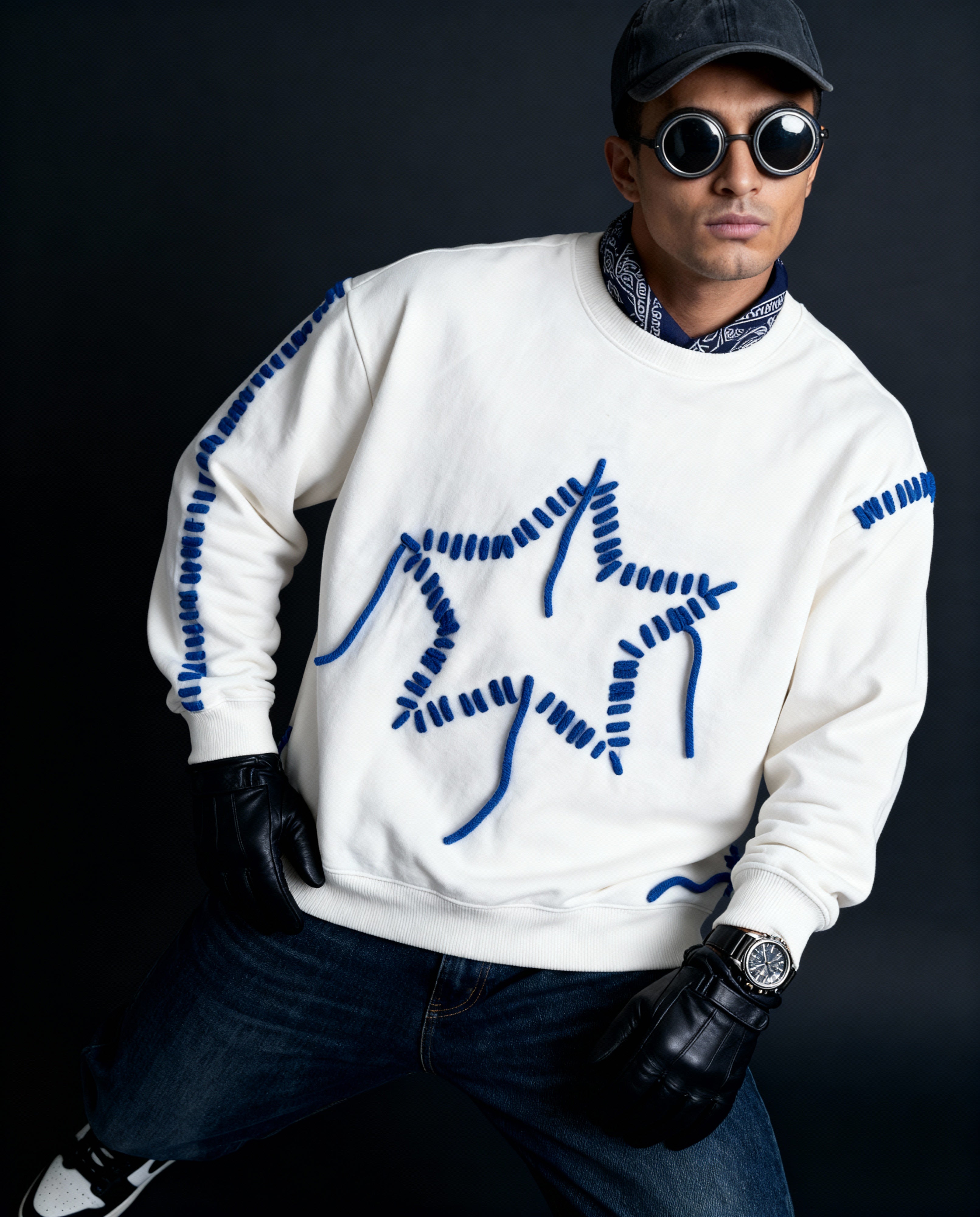 Person wearing a white sweater with blue embroidered designs, dark jeans, and black gloves against a dark background.