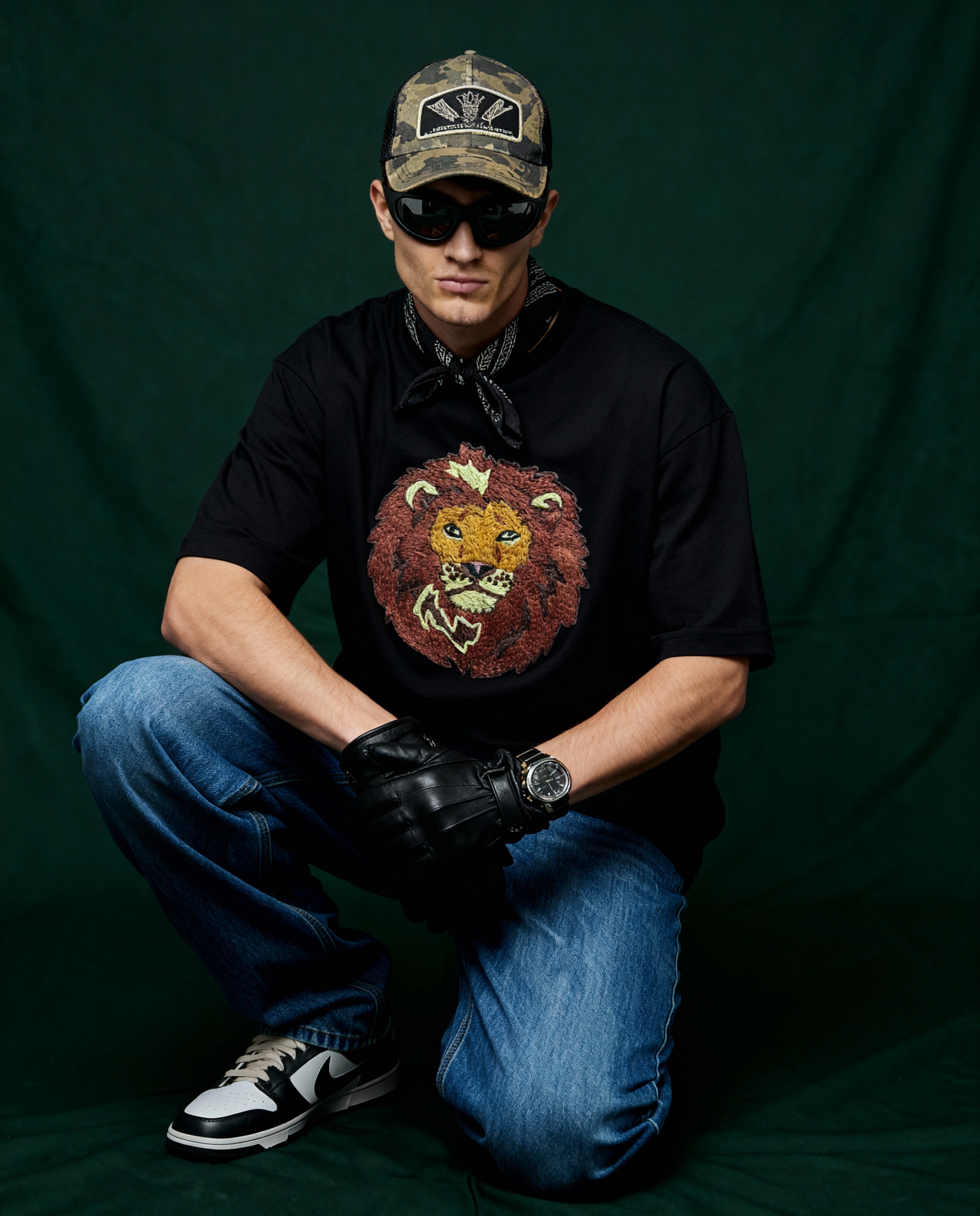Person wearing a black t-shirt with a lion embroidered, blue jeans, and a cap on a dark green background