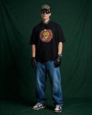 Person wearing a black t-shirt with a lion embroidery, blue jeans, black gloves, and sunglasses against a green backdrop.