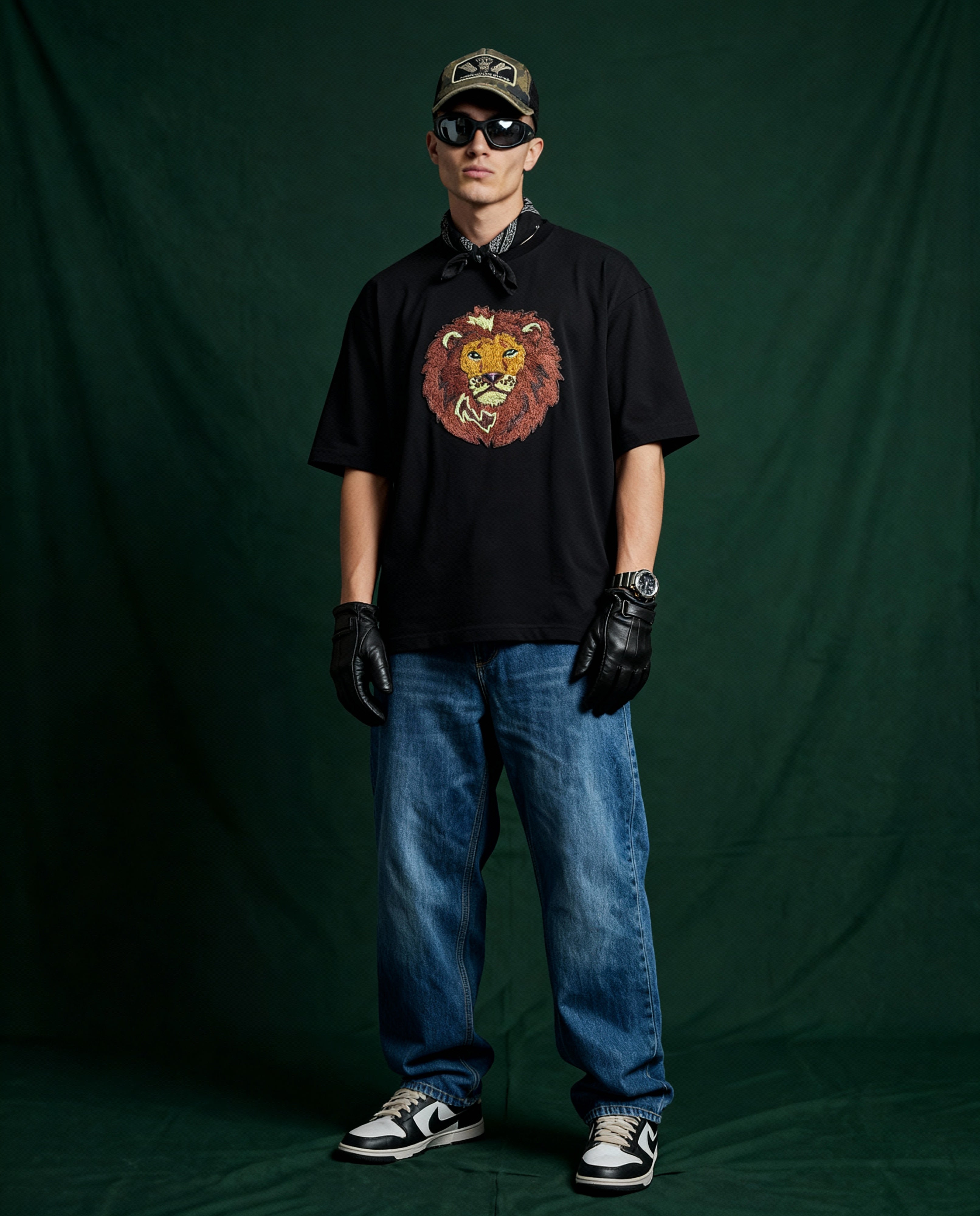 Person wearing a black t-shirt with a lion embroidery, blue jeans, black gloves, and sunglasses against a green backdrop.