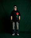 Person wearing a black t-shirt with a red rose design embroidered , standing against a dark green backdrop.