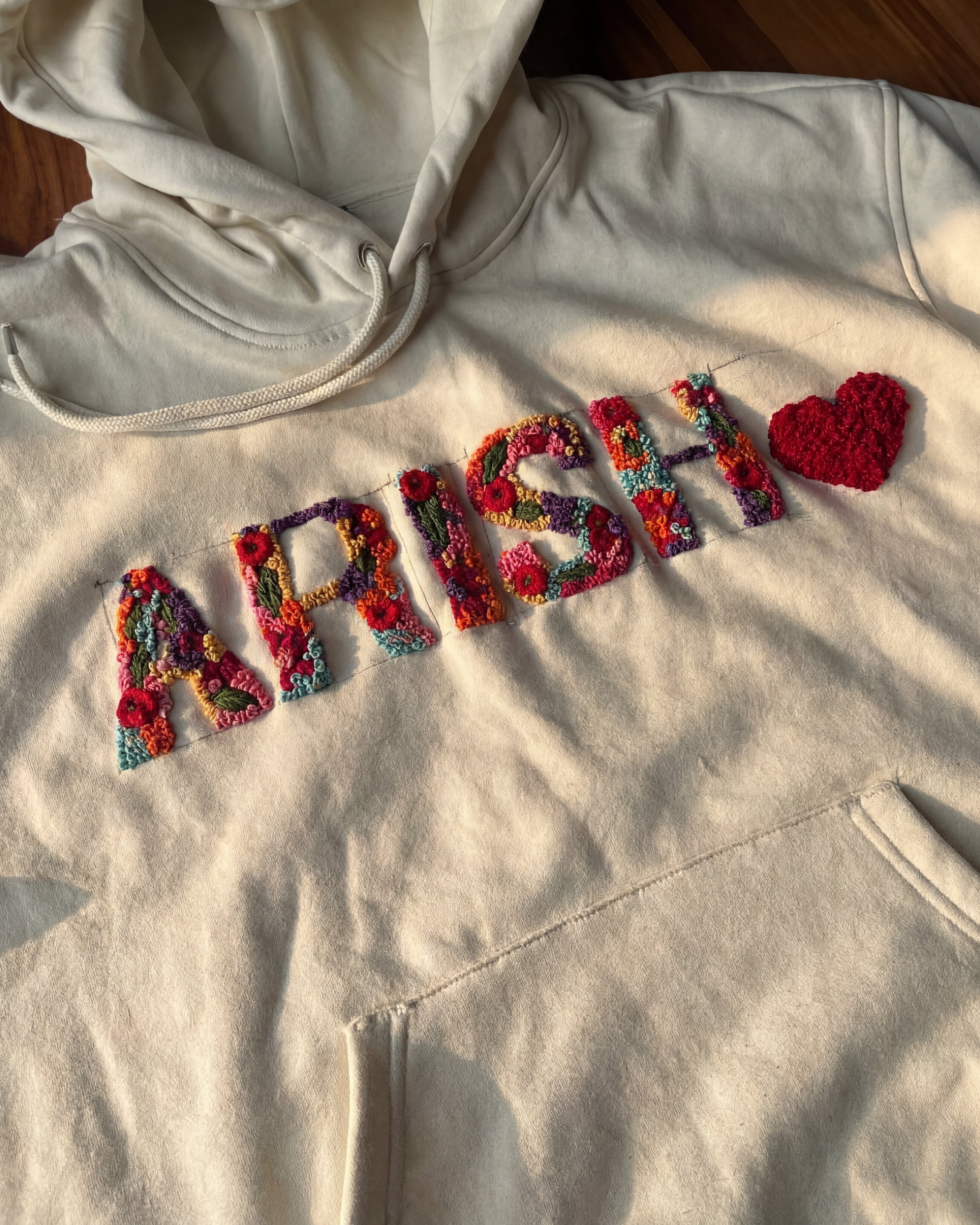 Beige hoodie with colorful customised embroidered letters spelling 'APRIL' on a wooden surface