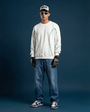 Person wearing a white sweater with patterned embroidered sleeves, blue jeans, black gloves, and a cap on a dark background