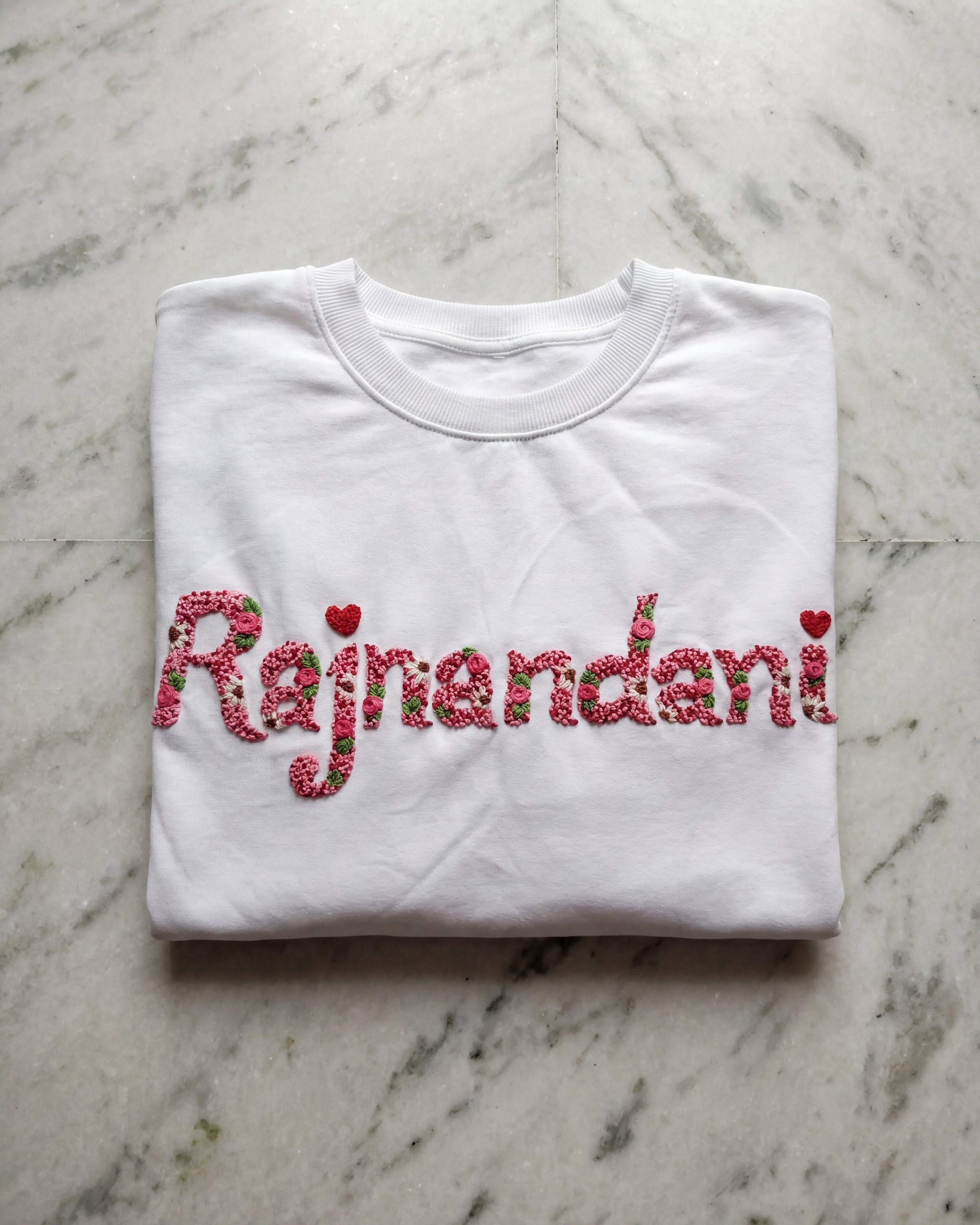 Folded white t-shirt with 'Rajnandan' embroidered in red on a marble surface