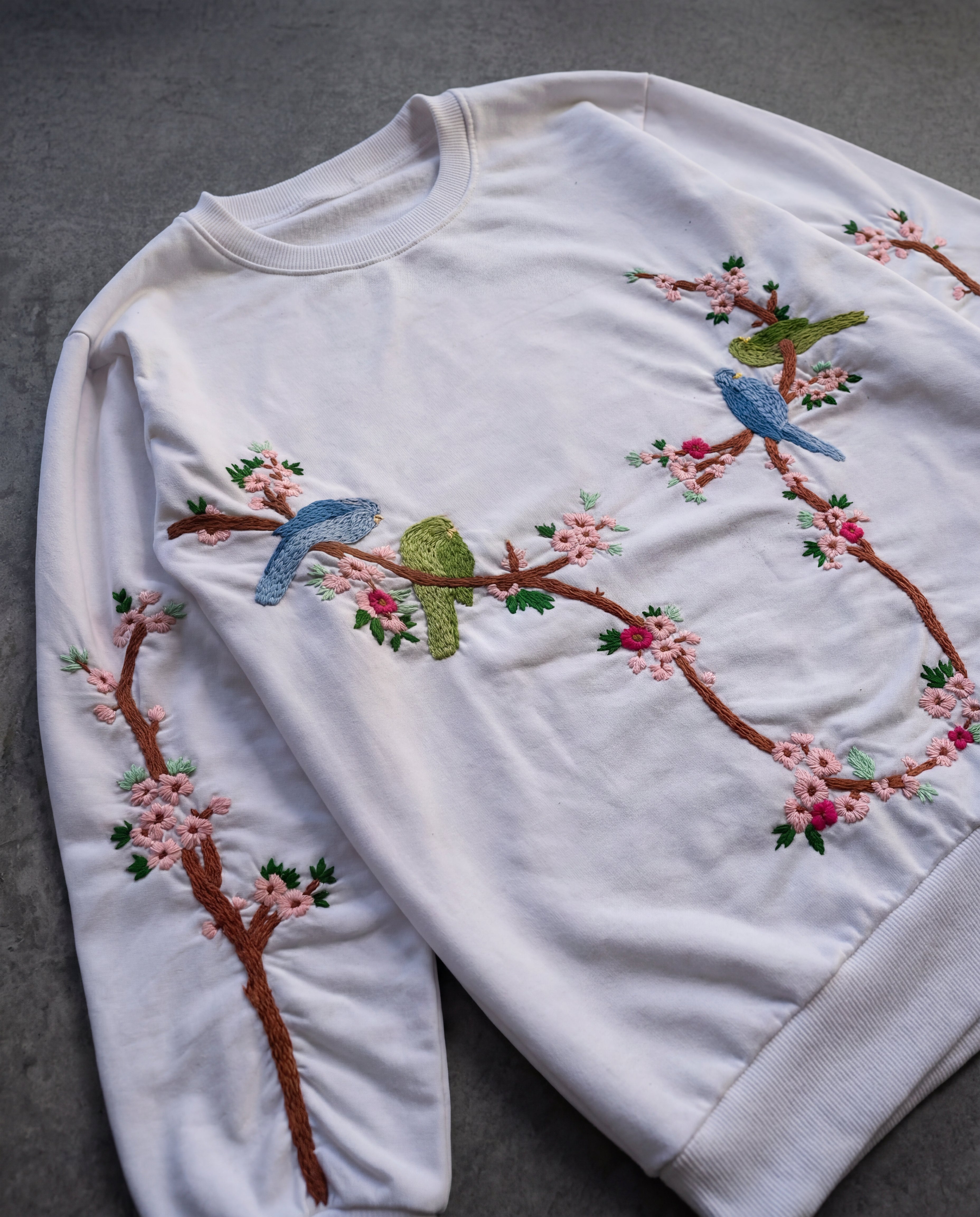 White sweatshirt with embroidered birds and cherry blossoms on a gray background