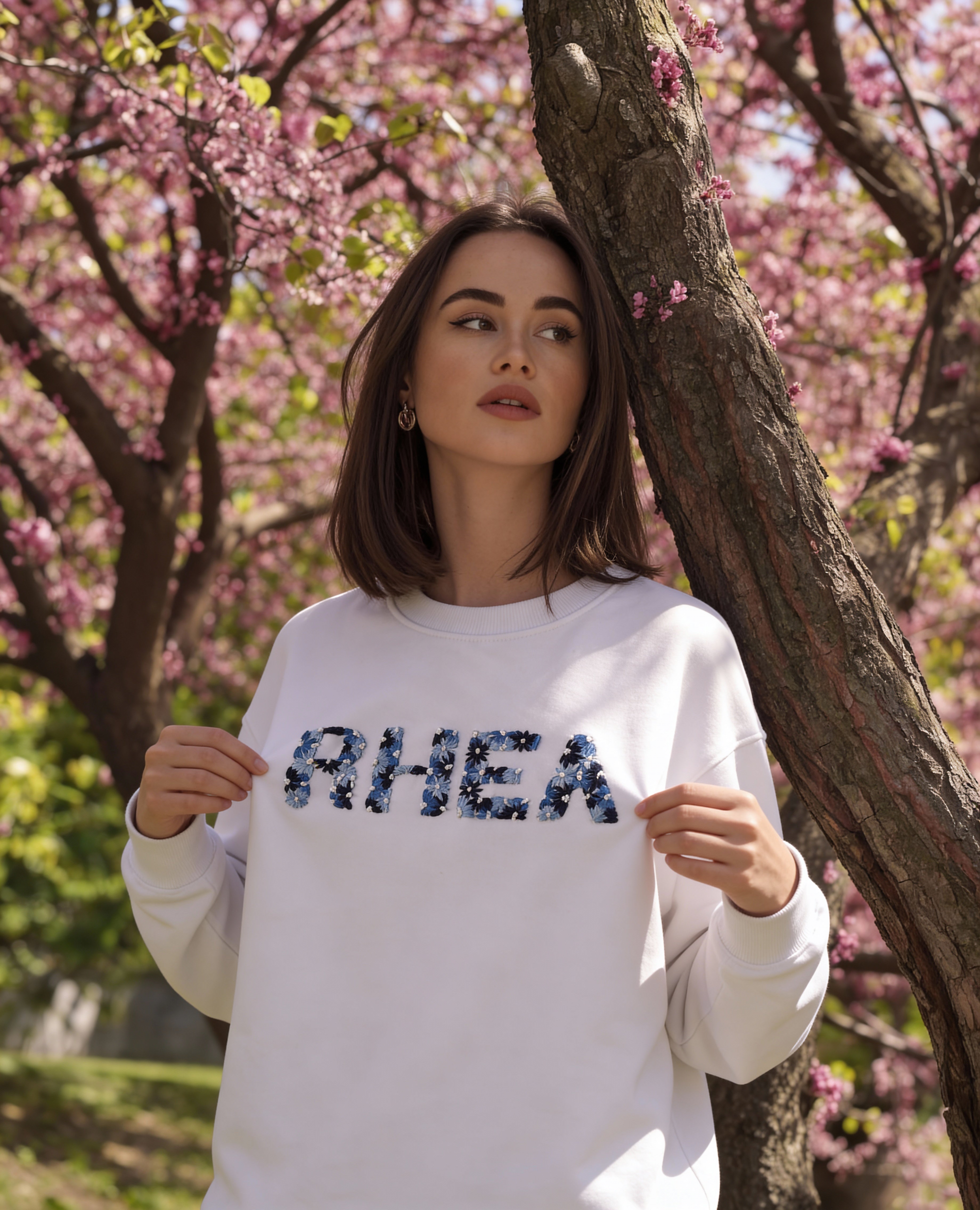 Woman wearing a white sweatshirt with customised name hand embroidered  'RIHEA'  on it, standing in a cherry blossom garden