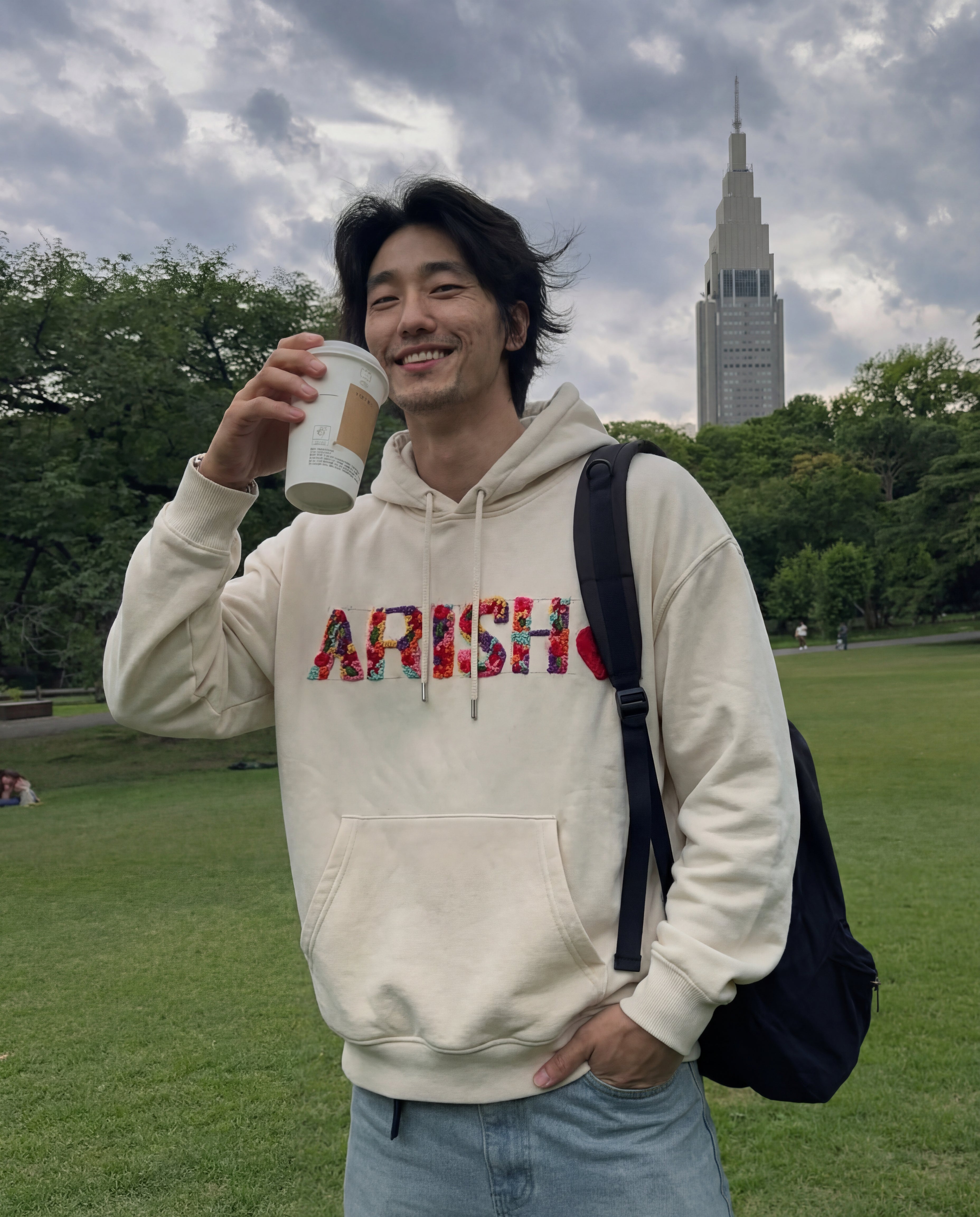 Man in a beige hoodie with 'ARISH' holding a coffee cup outdoors with a tall building in the background.