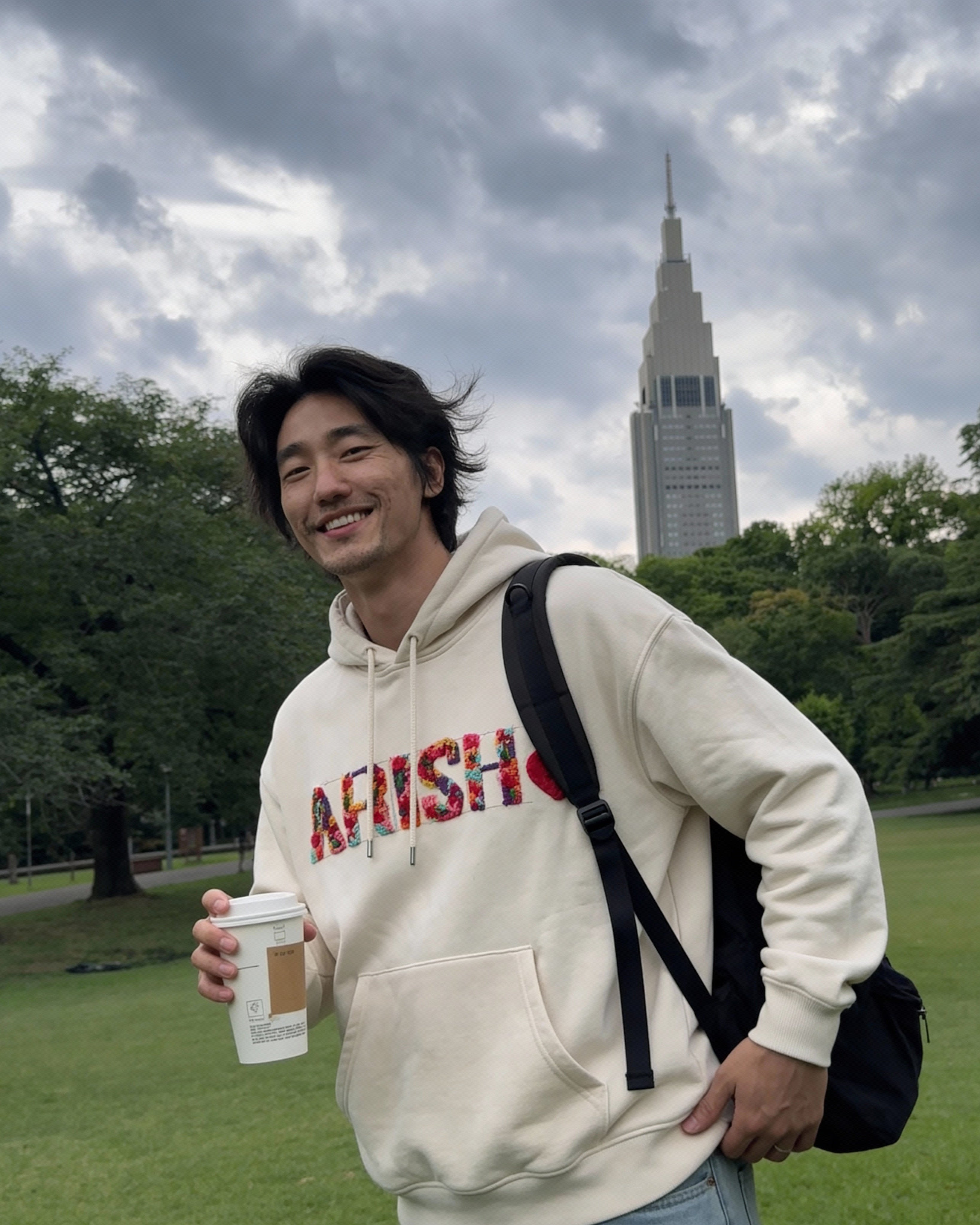 Man in a hoodie with a visible brand logo, holding a coffee cup, standing in a park with a tall building in the background.