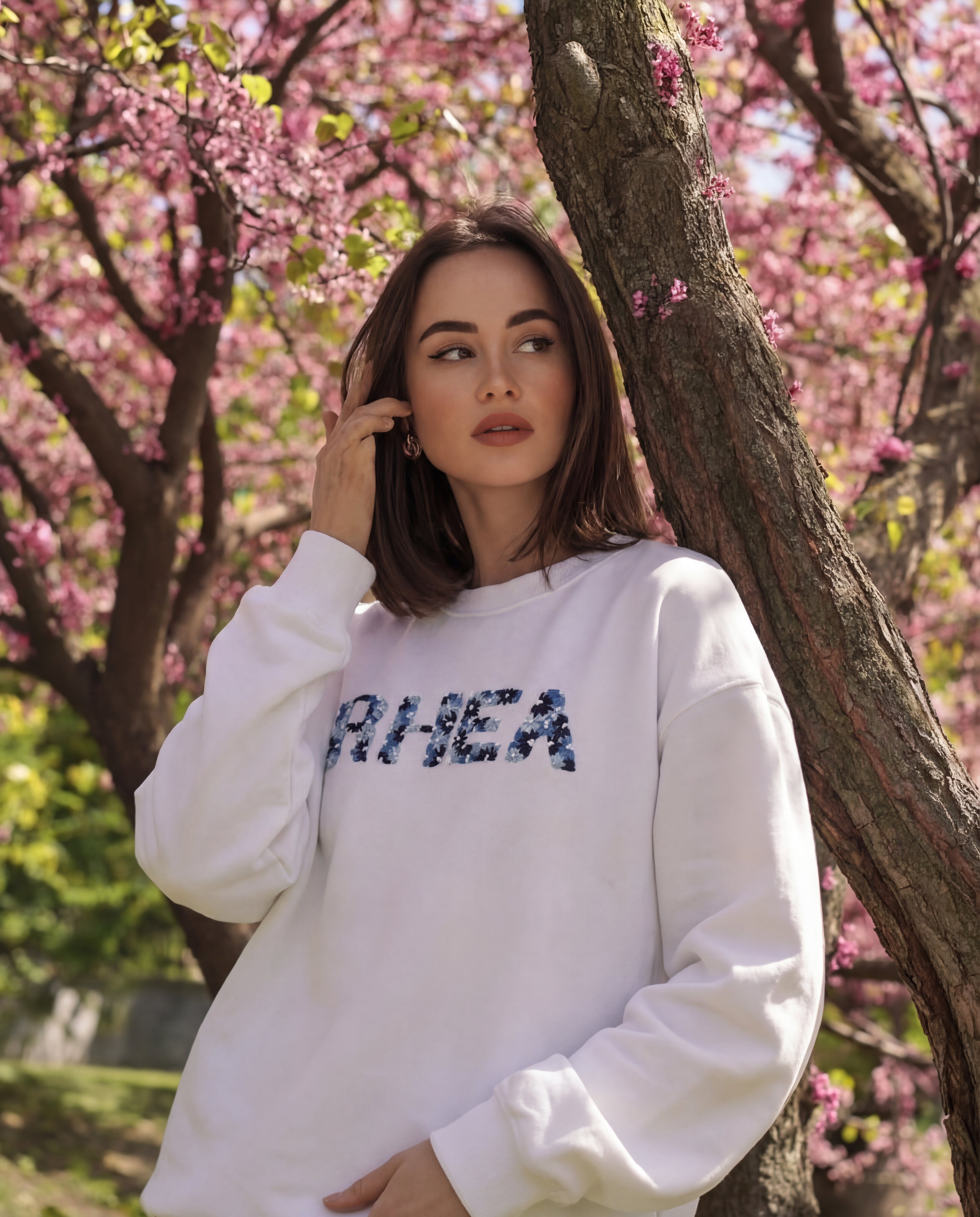 Woman wearing a white sweatshirt with name  'RHEA' embroidered  on it, standing in front of cherry blossom trees.
