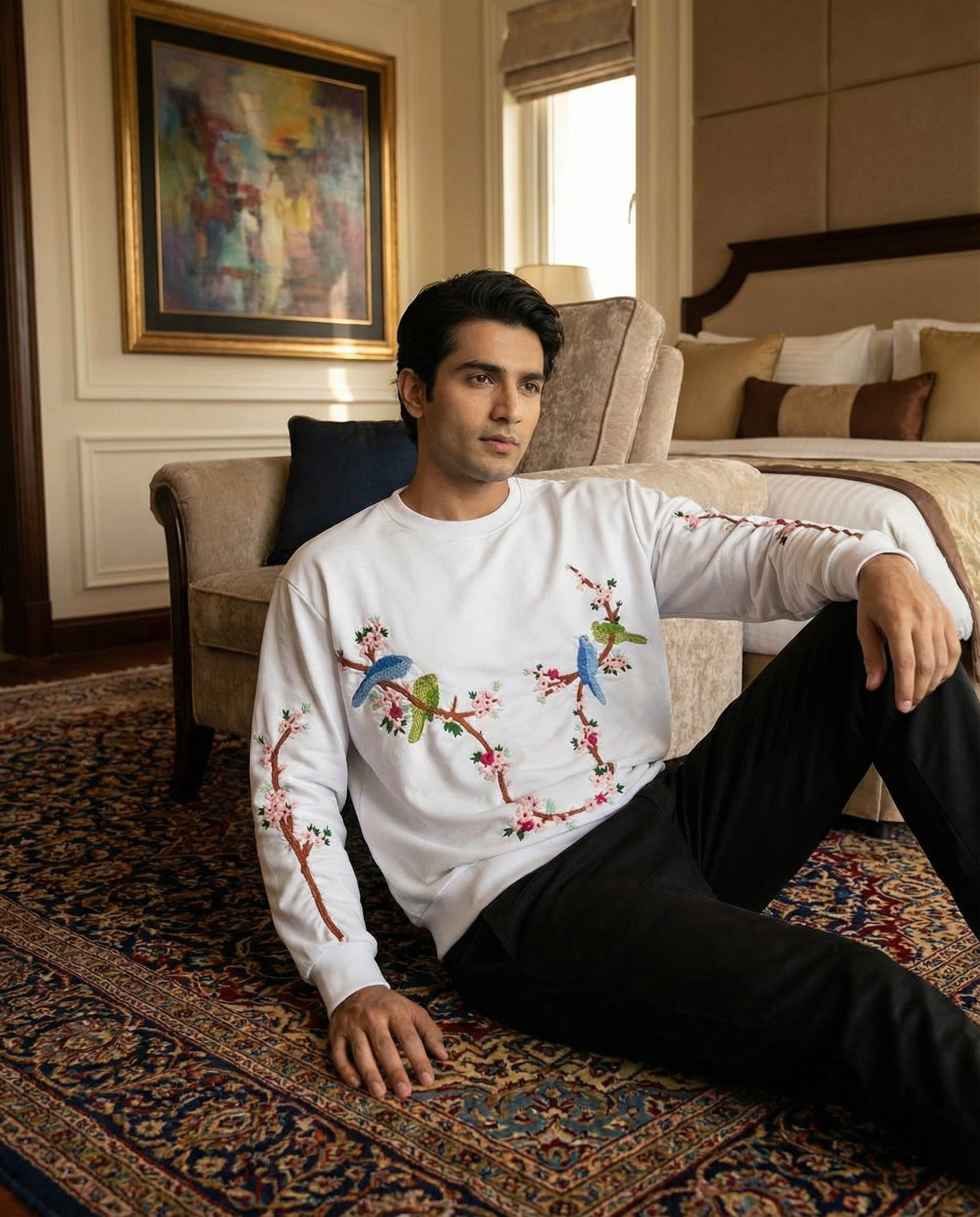Man wearing a white sweater with colorful bird embroidered design sitting on a couch in a living room.