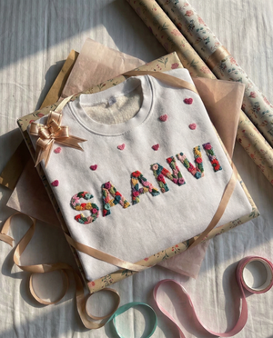 Sweatshirt with colorful custom name embroidery  on a wooden surface with ribbons and paper.