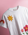 White t-shirt with embroidered sun, rainbow, and flower designs on a pink background