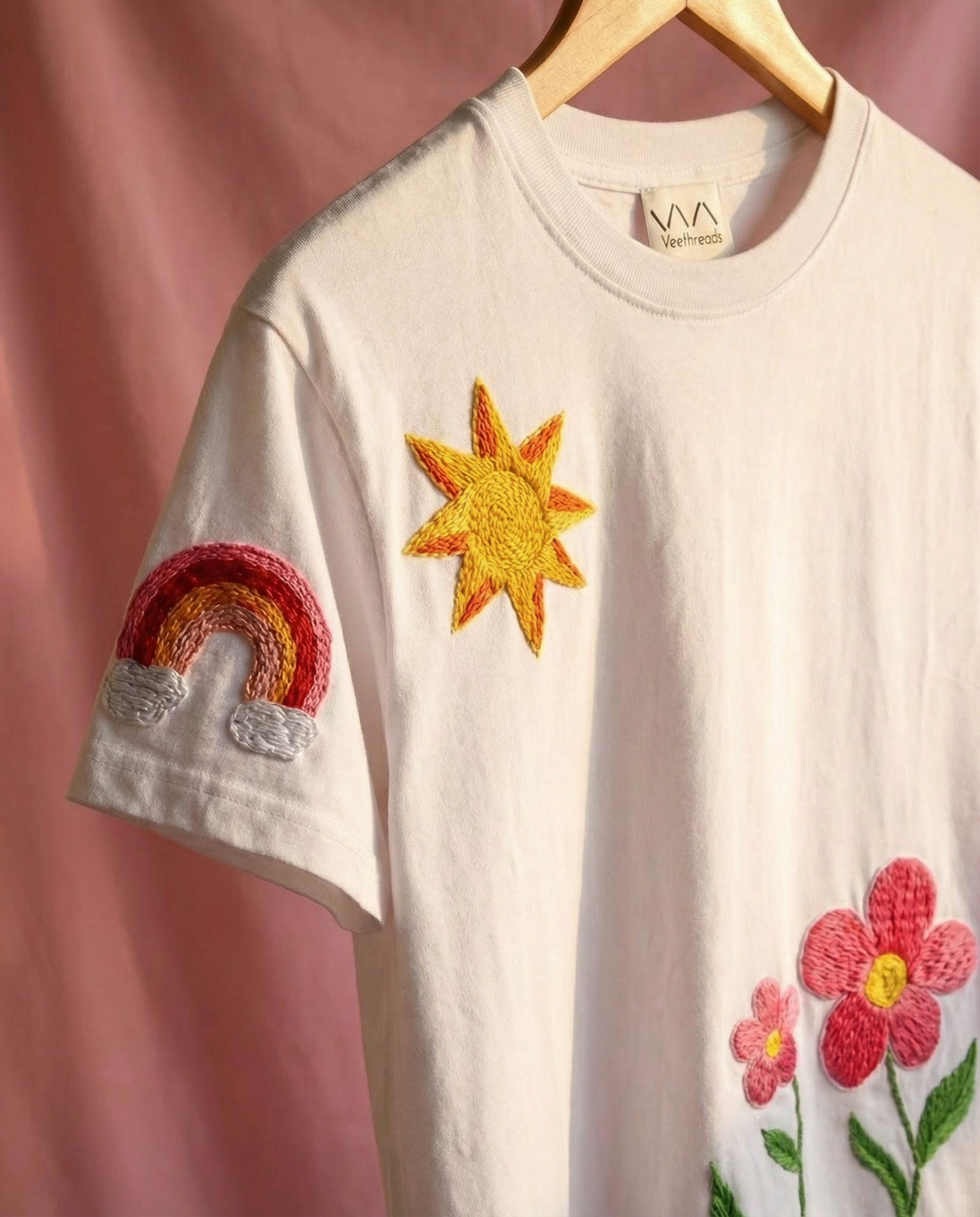 White t-shirt with embroidered sun, rainbow, and flower designs on a pink background