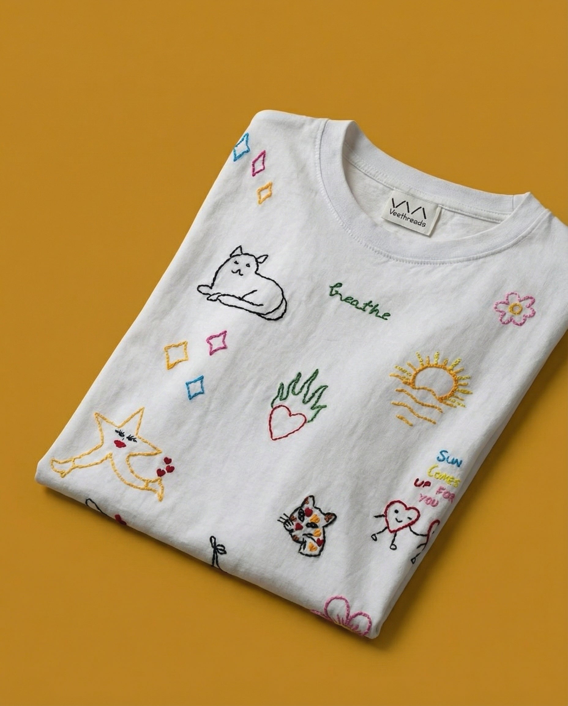 Embroidered doodle tee flatlay — folded white tee with colourful hand-embroidered doodle designs on mustard yellow background